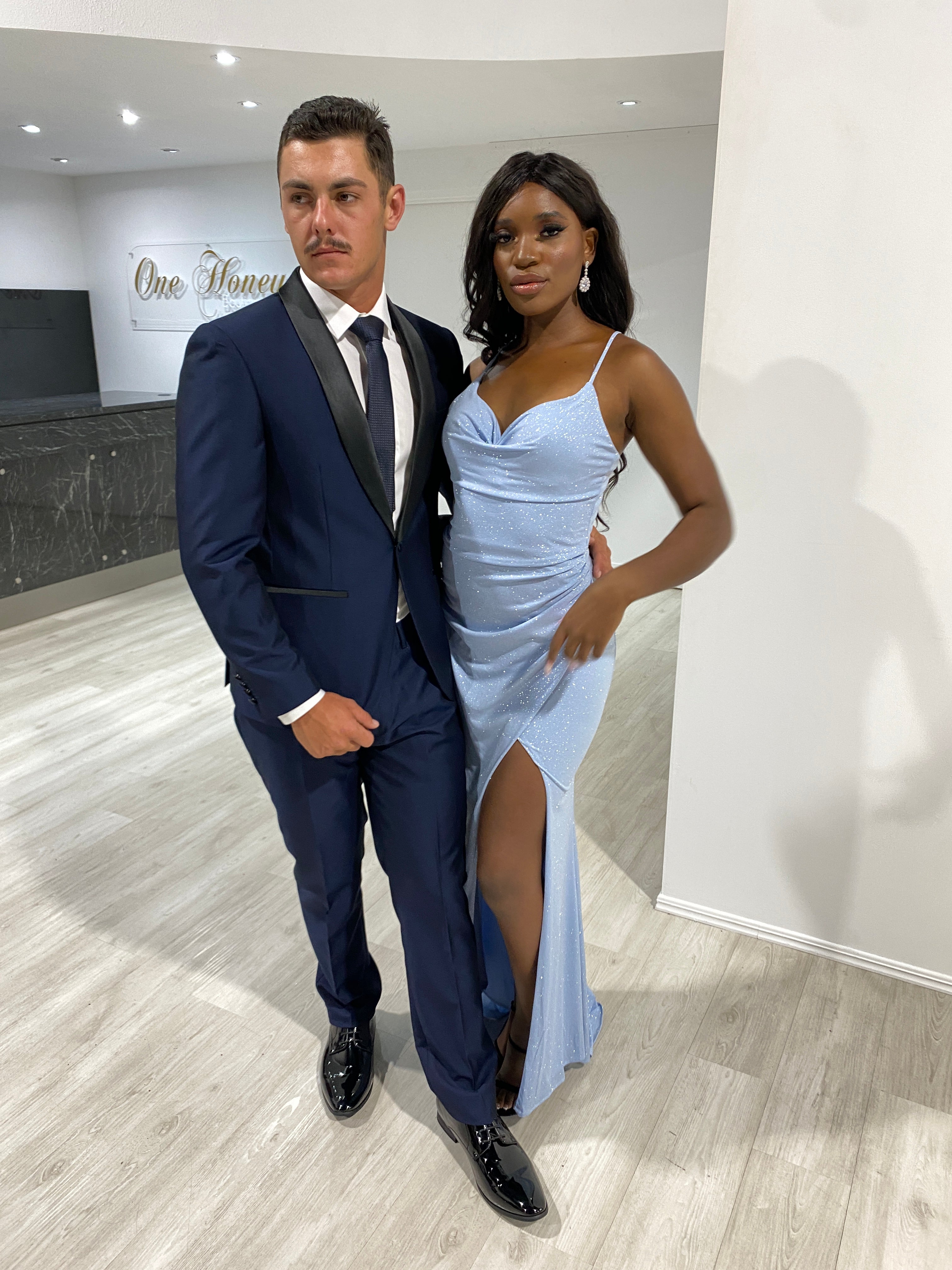 Elegant couple in formal attire, featuring a blue mermaid gown with a thigh-high slit and a sleek blue suit, posing at an event.
