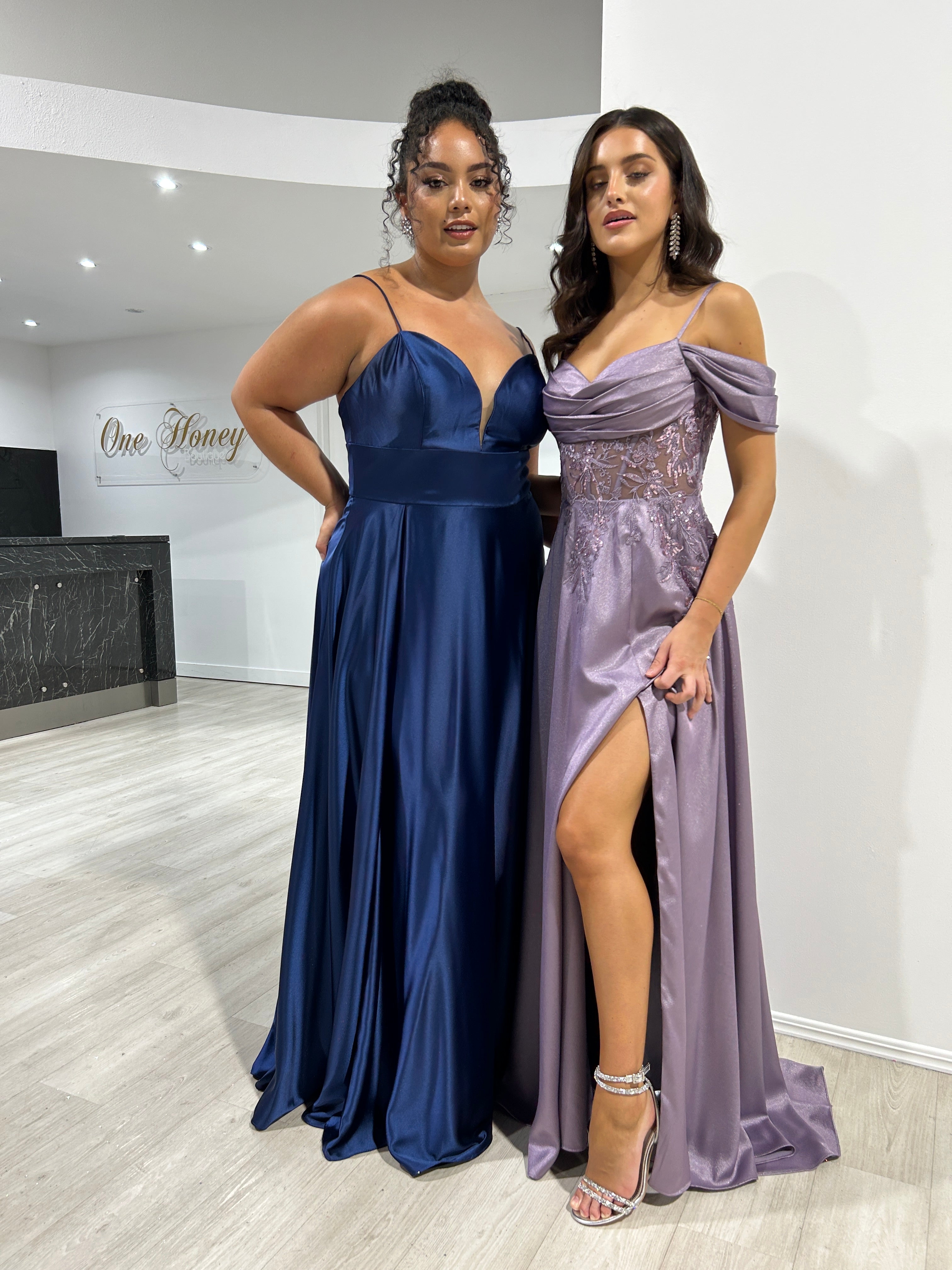 Two women in elegant gowns, one in a navy satin dress and the other in a lilac beaded off-shoulder dress with a thigh-high slit, Brisbane to Gold Coast.