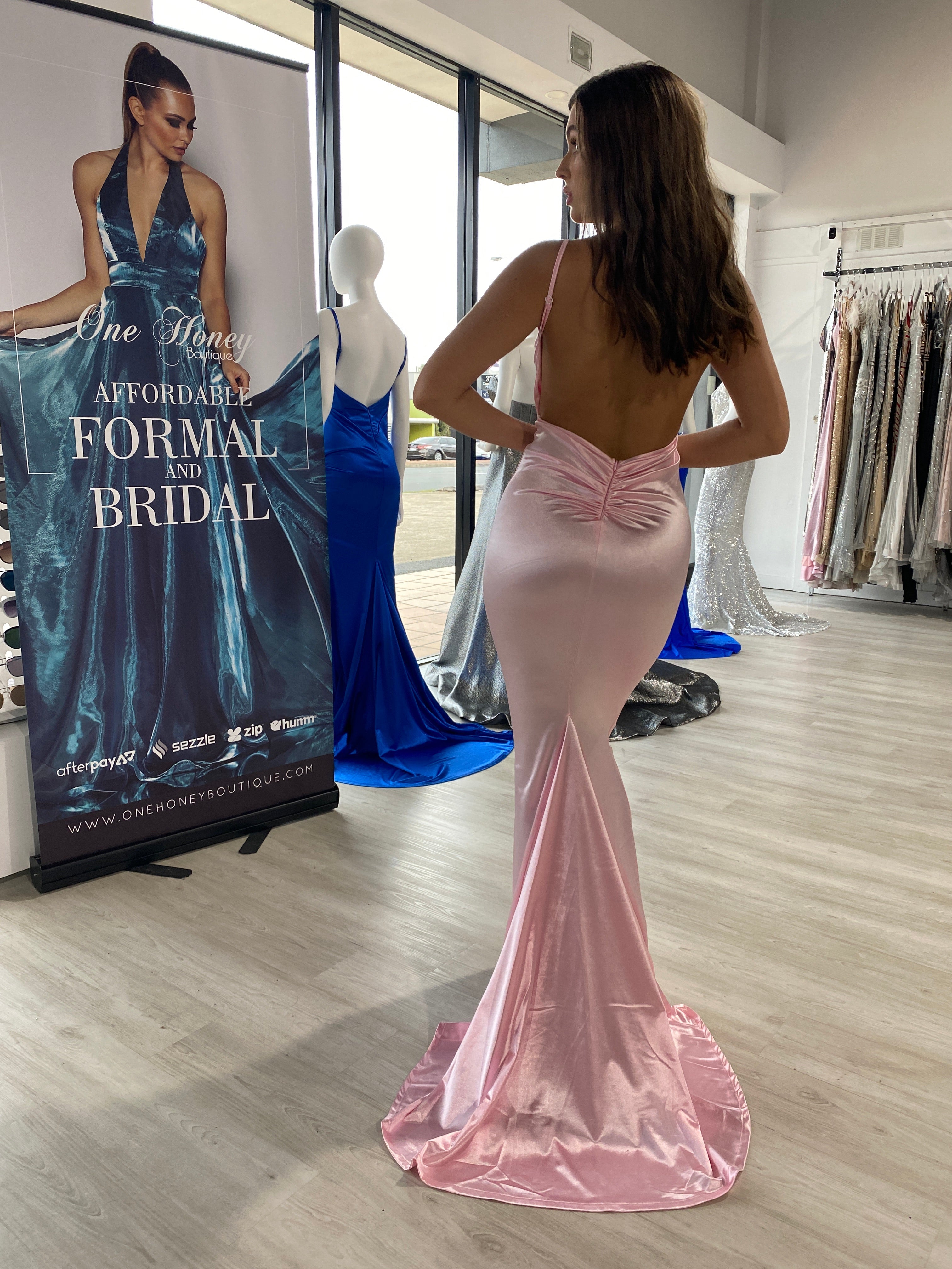 Honey Couture MILEE Low Back Mermaid Evening Gown in silky pink fabric displayed in boutique, highlighting bum ruching and elegant design.