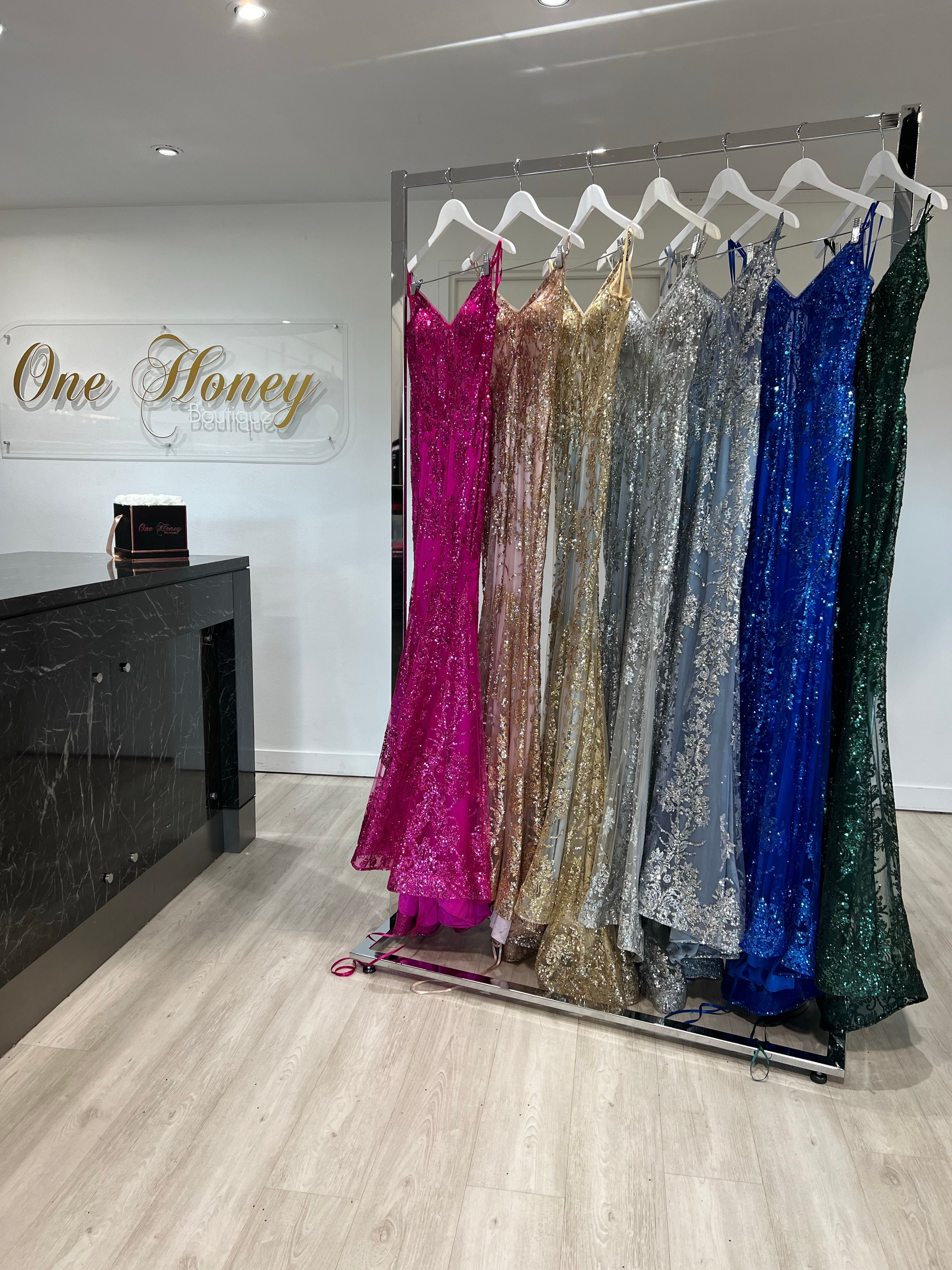Various glittering evening gowns, including sequin and embroidered dresses, displayed on hangers in a boutique. One Honey signage visible.