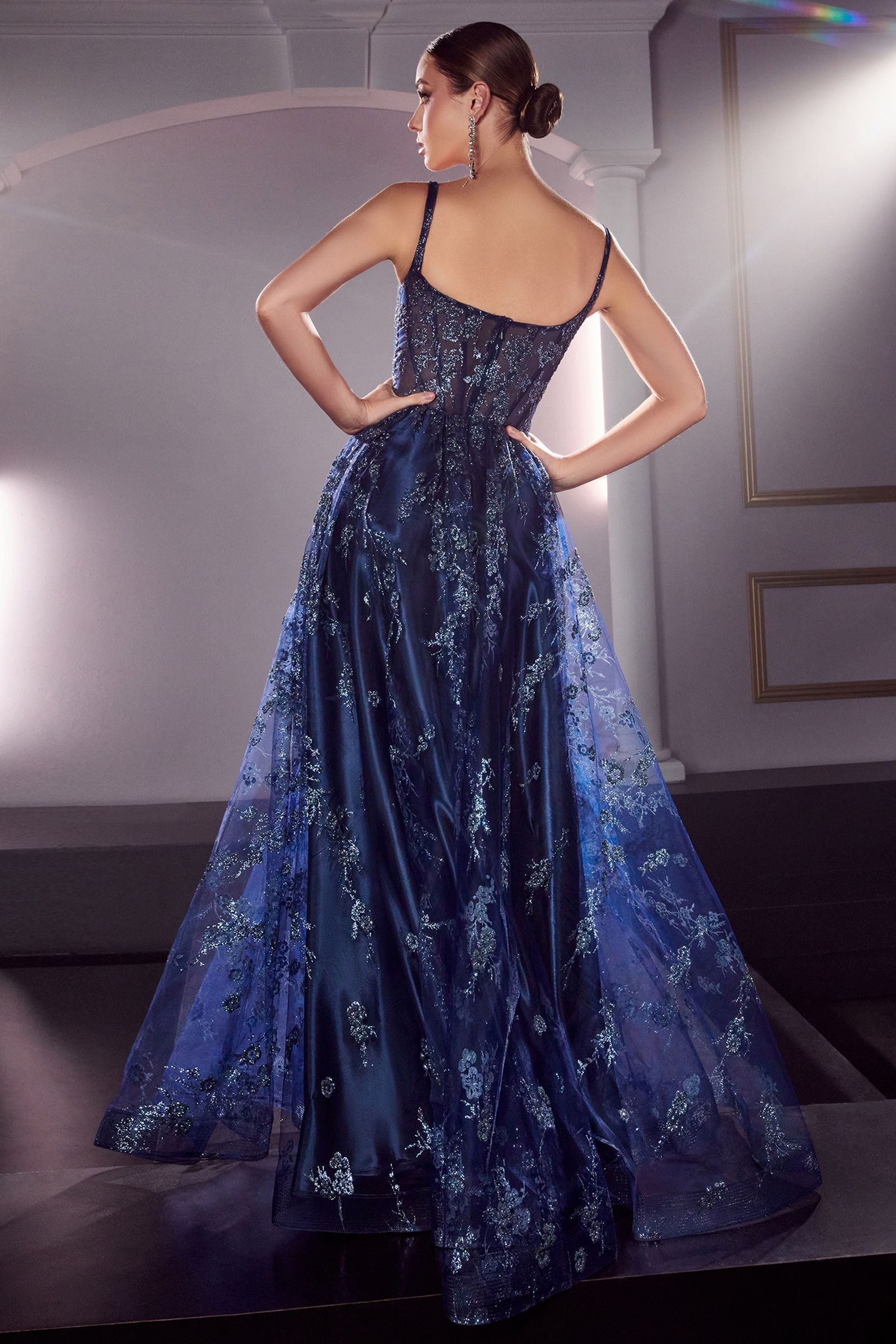 A woman in a navy blue lace ball gown with floral embroidery, featuring a fitted corset and flowing skirt. Formal setting.