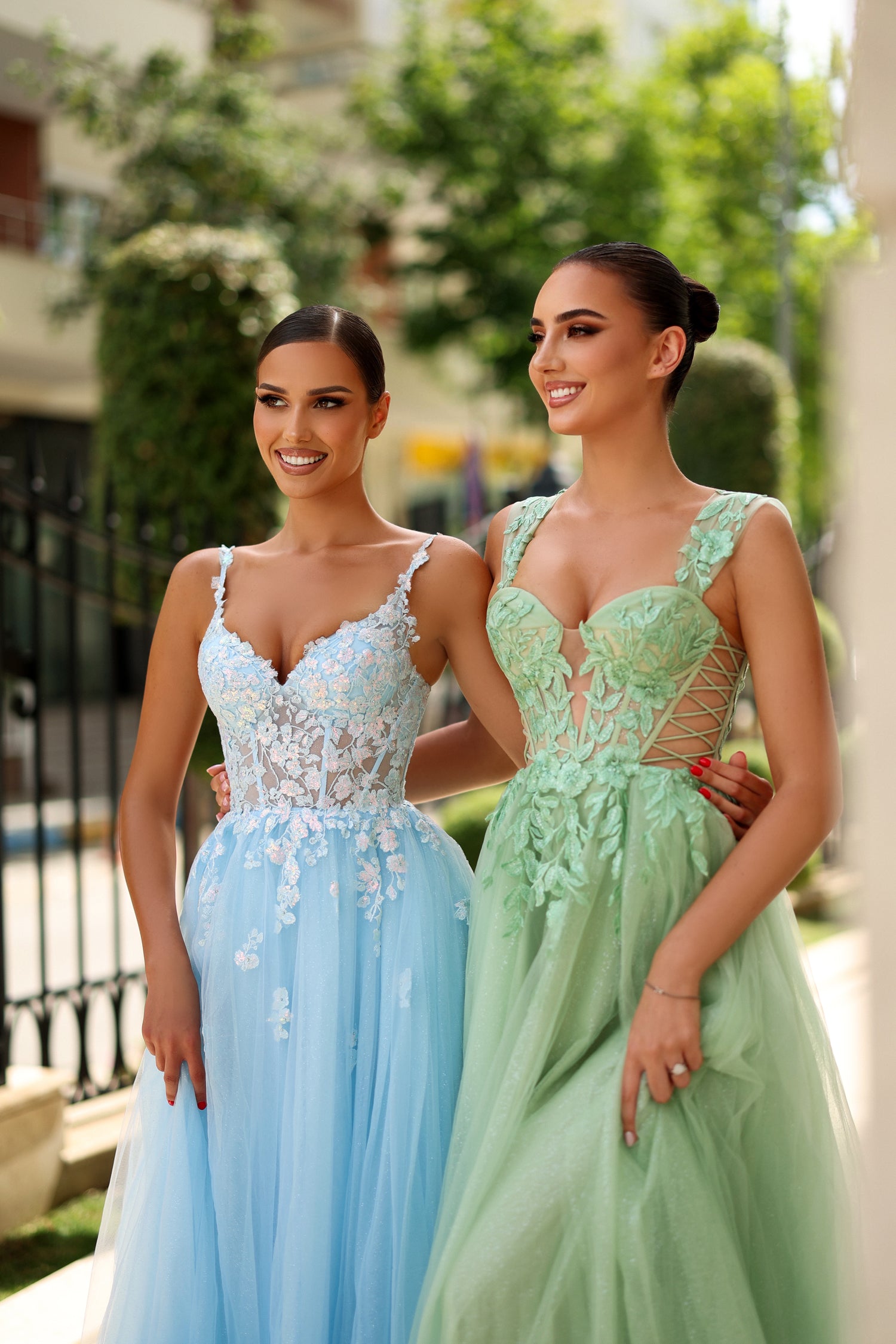 Two women in elegant lace tulle prom gowns, one in blue and the other in green, with floral appliqué details, outdoors on a sunny day.