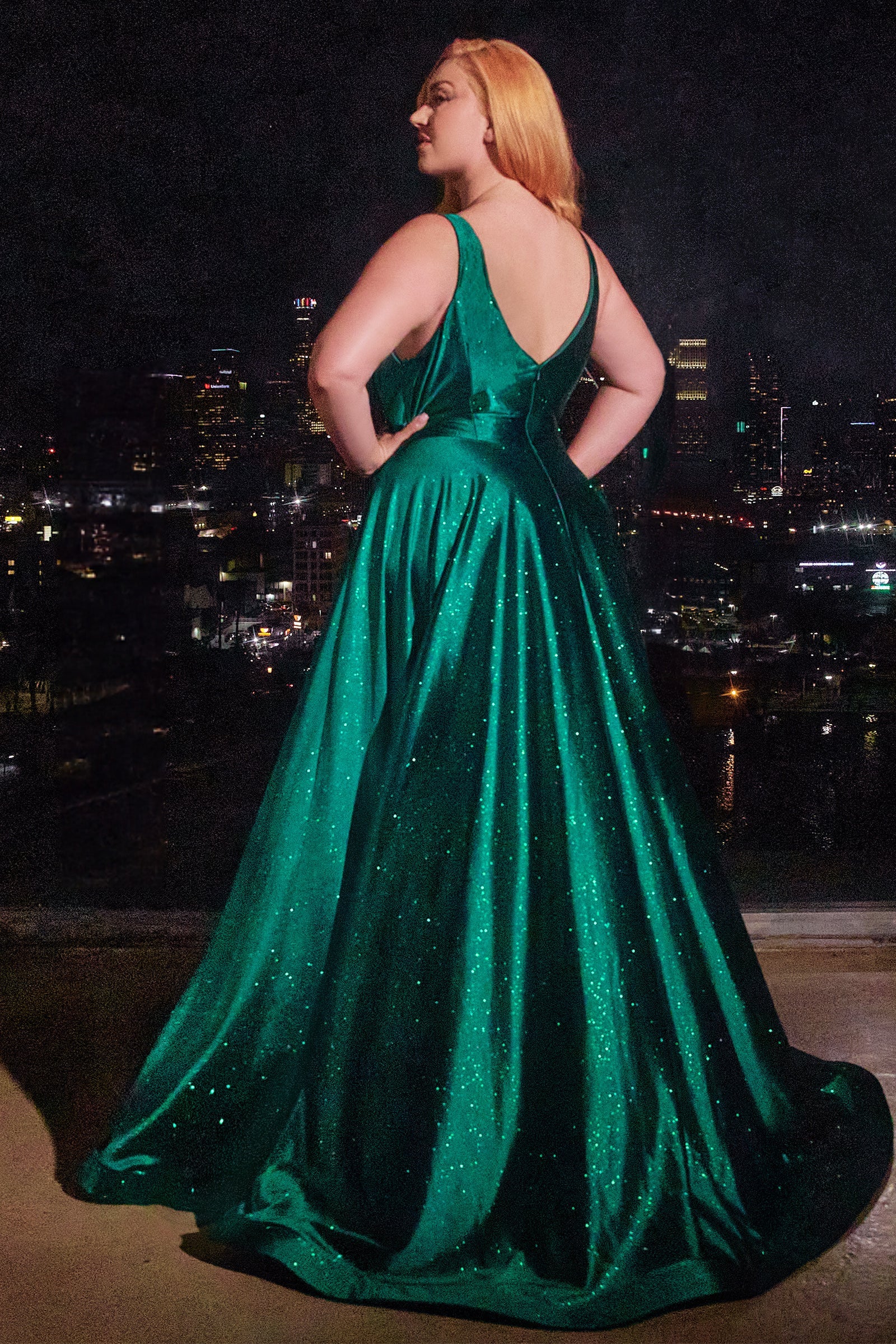 Plus size woman in a shimmering green A-line dress, featuring a deep V-back, looking out at the city skyline at night.