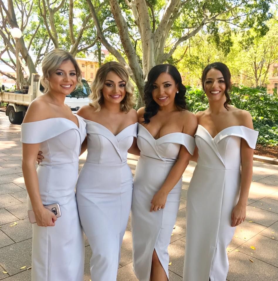 Four women in elegant off-shoulder white midi dresses with side slits, standing outdoors under trees, smiling for a group photo.