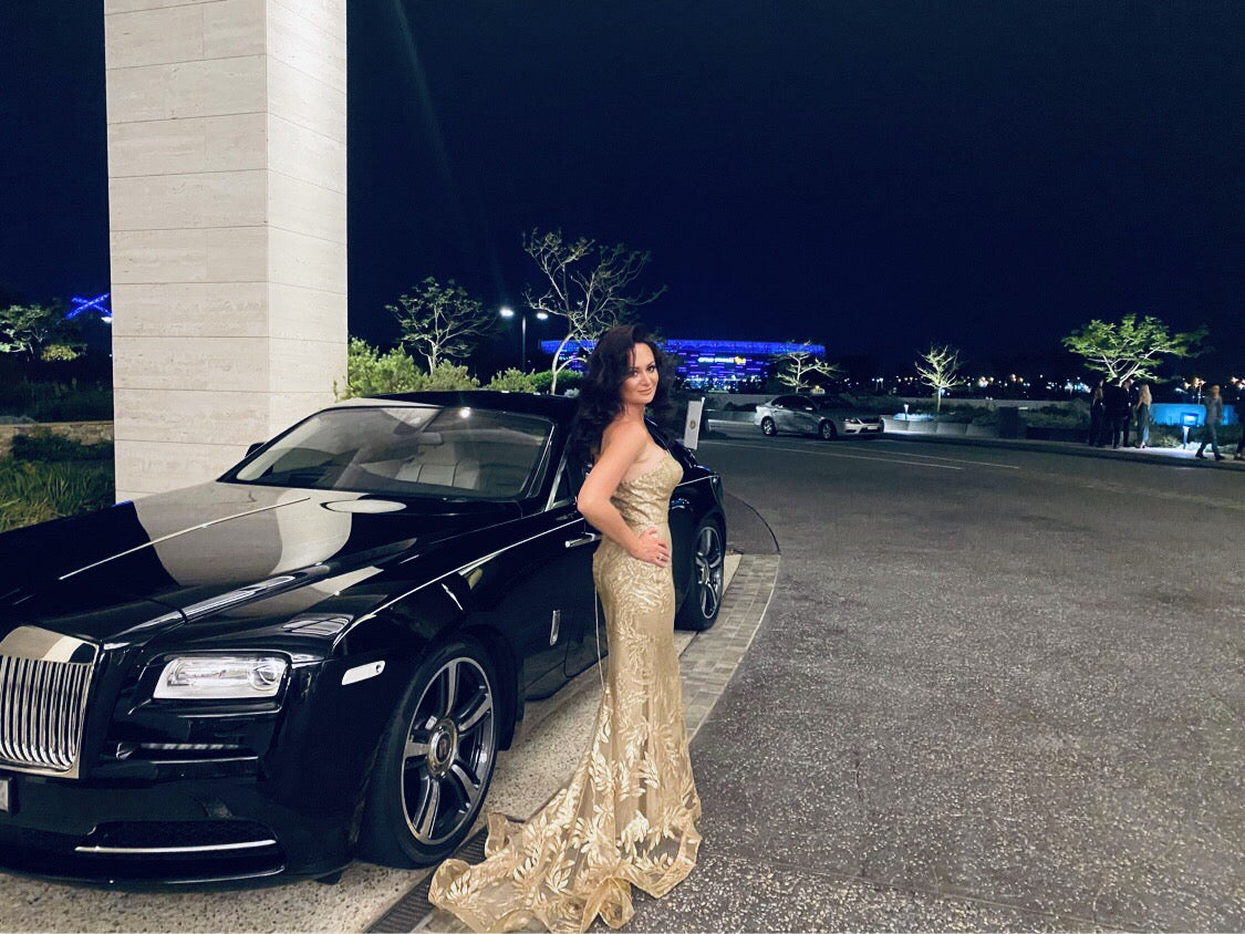Woman in a gold strapless mermaid formal dress standing beside a luxury black car at night, showcasing elegance and glamour.