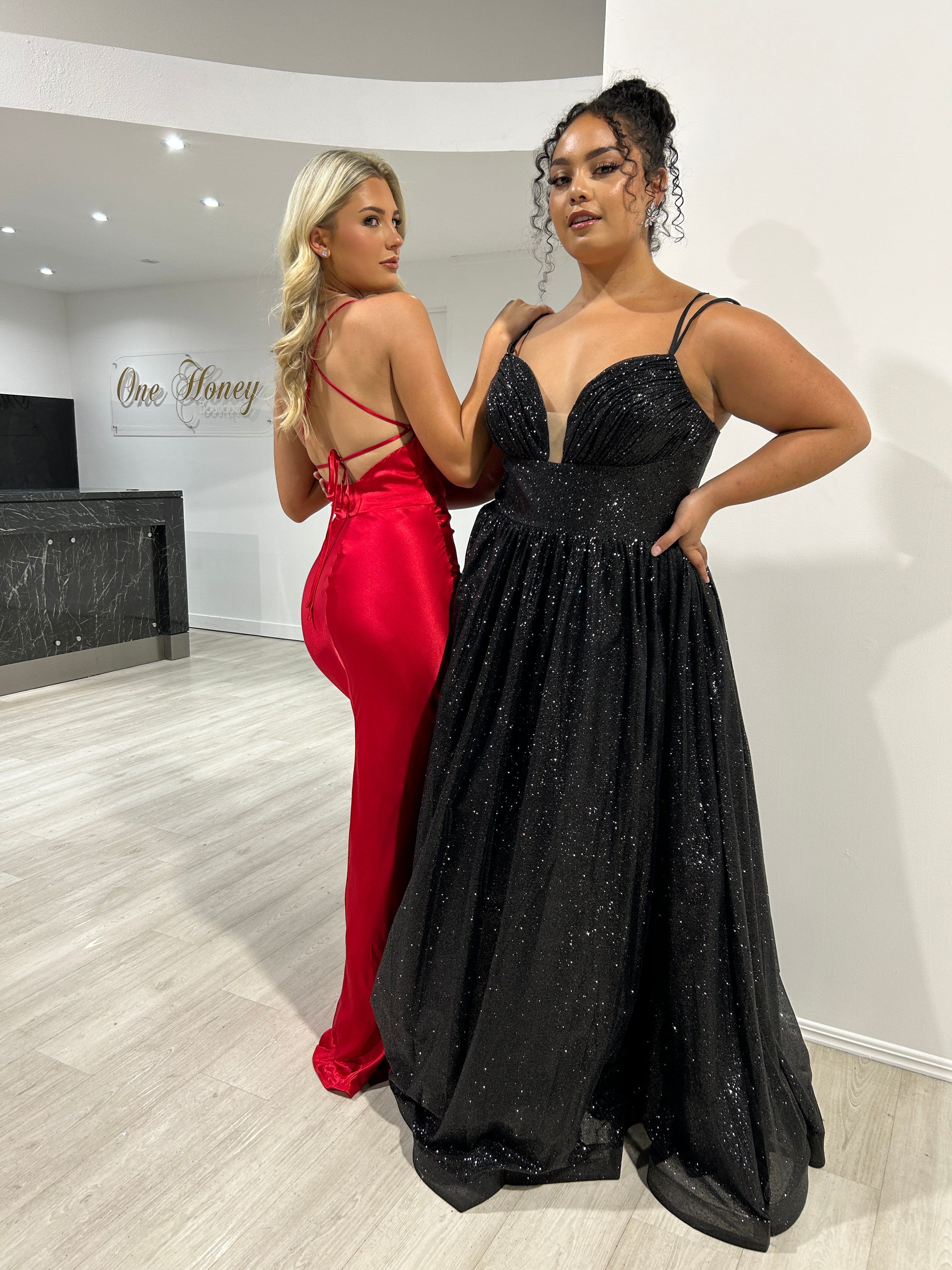 Two women in elegant evening gowns, one in a red satin dress with a lace-up back, the other in a black glitter gown. Brisbane to Gold Coast.