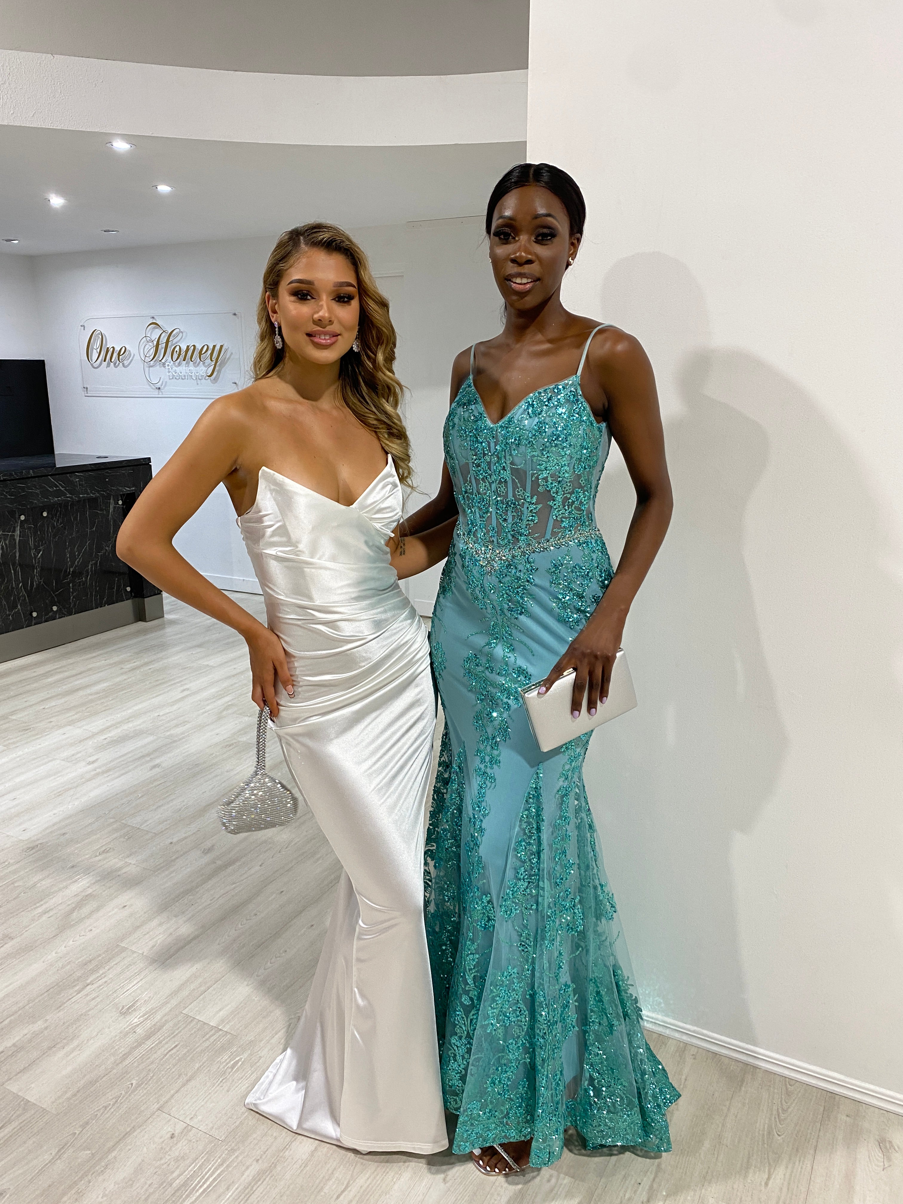 Two women wearing elegant prom dresses, one in a white satin strapless gown and the other in a turquoise lace dress, at One Honey.