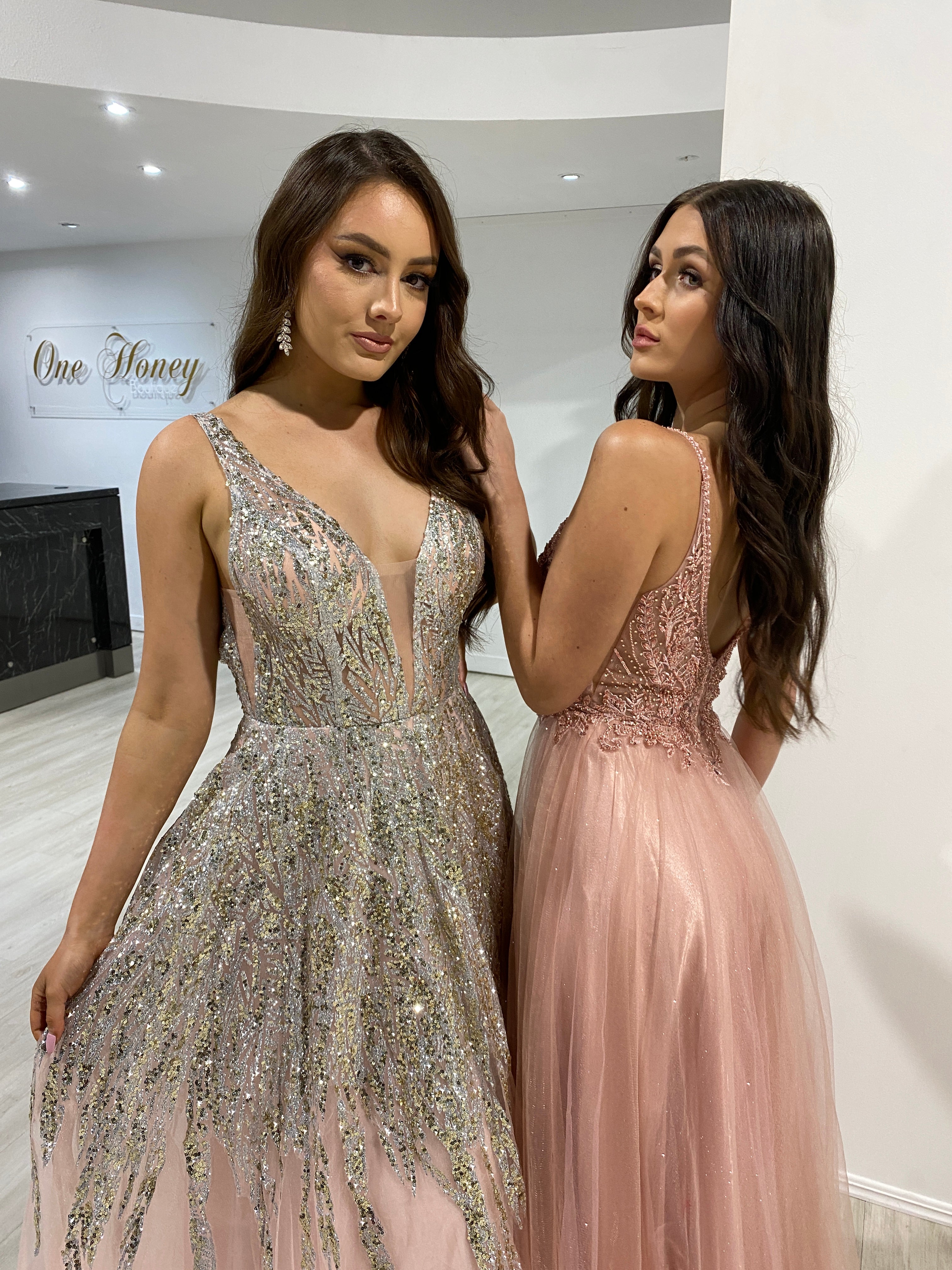 Two women in elegant prom gowns, one with a glittering bodice and the other with a lace appliqué, standing in a modern boutique.