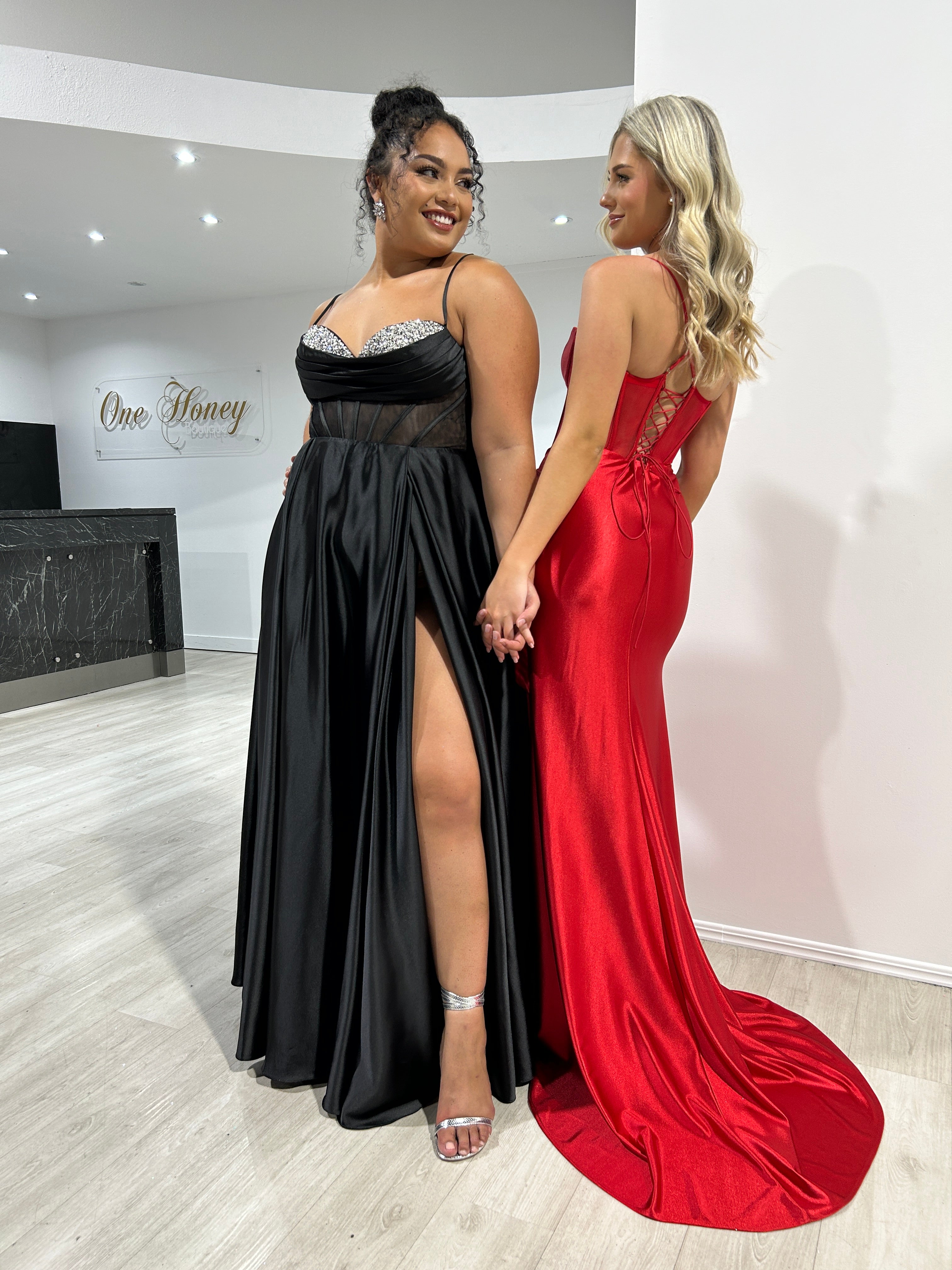 Two women in elegant formal gowns, one in a black lace slit dress and one in a red satin mermaid dress, standing side by side.