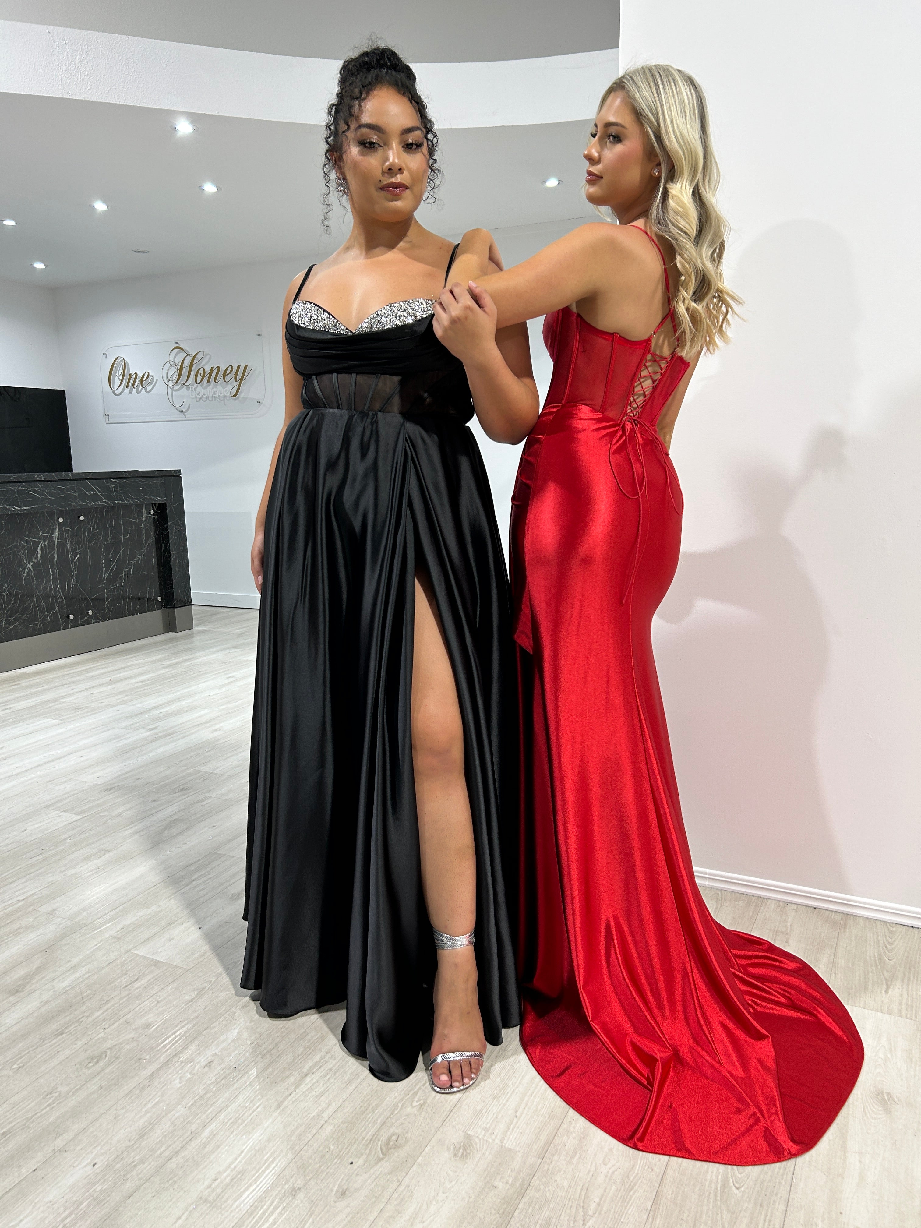 Two women in a boutique wearing elegant satin gowns, one black with a leg slit and one red with lace-up back, perfect for formal events.