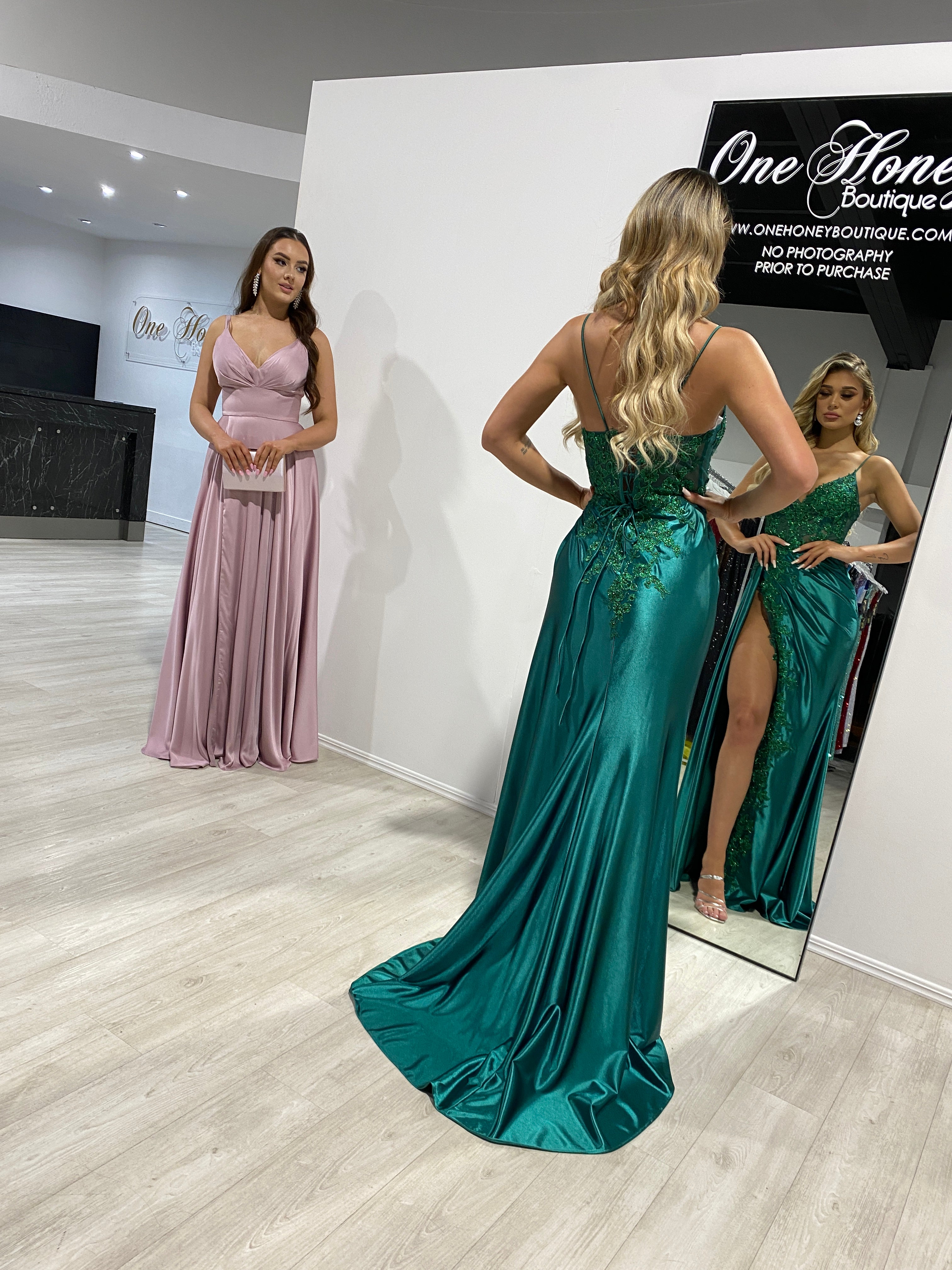 Two women in a dress boutique, one wearing a green satin gown with an open back and leg slit, the other in a pink A-line dress.