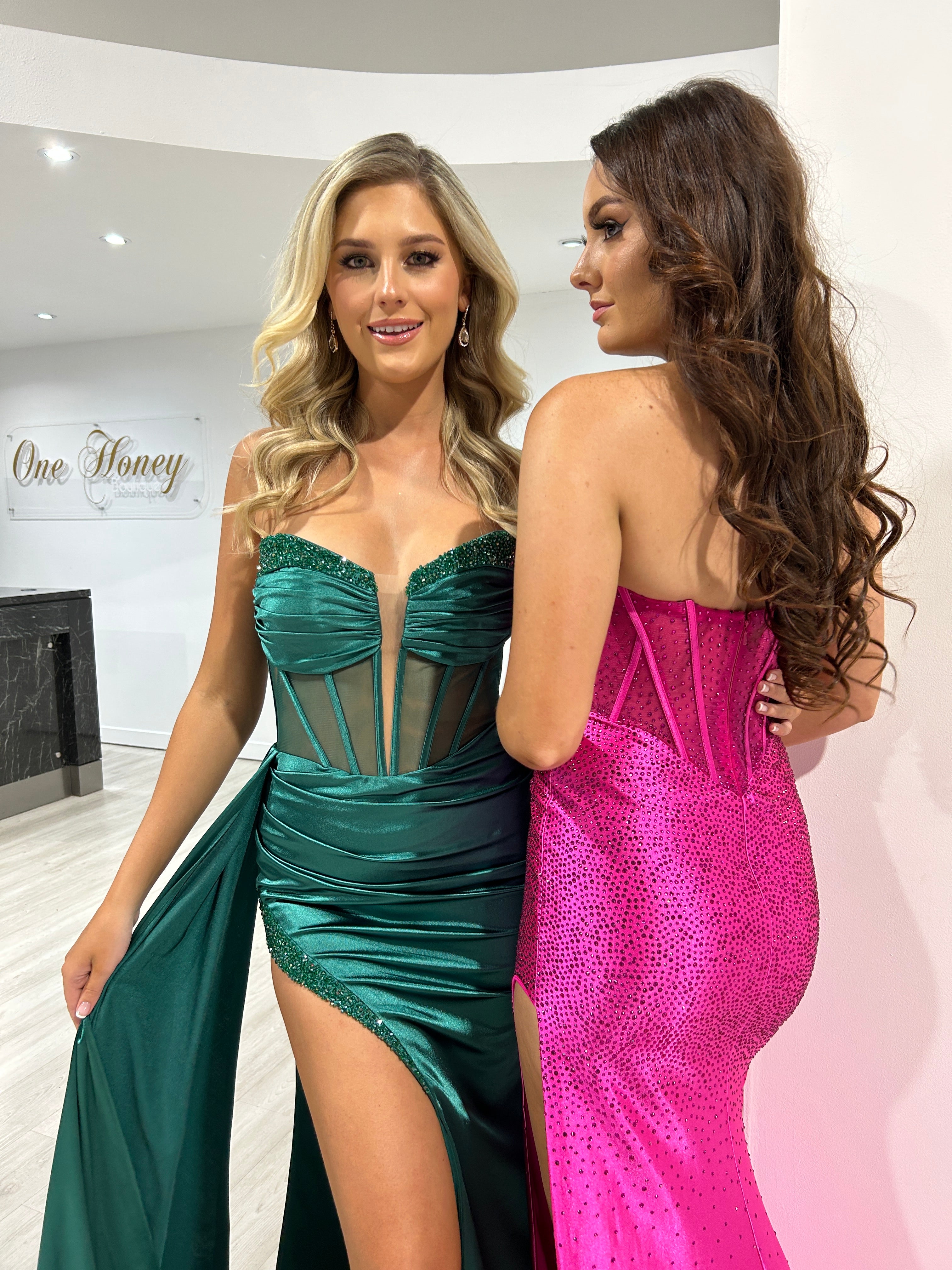 Two women wearing glamorous formal dresses, one in an emerald green corset gown with a high slit, and the other in a pink glitter corset dress. Brisbane to Gold Coast.