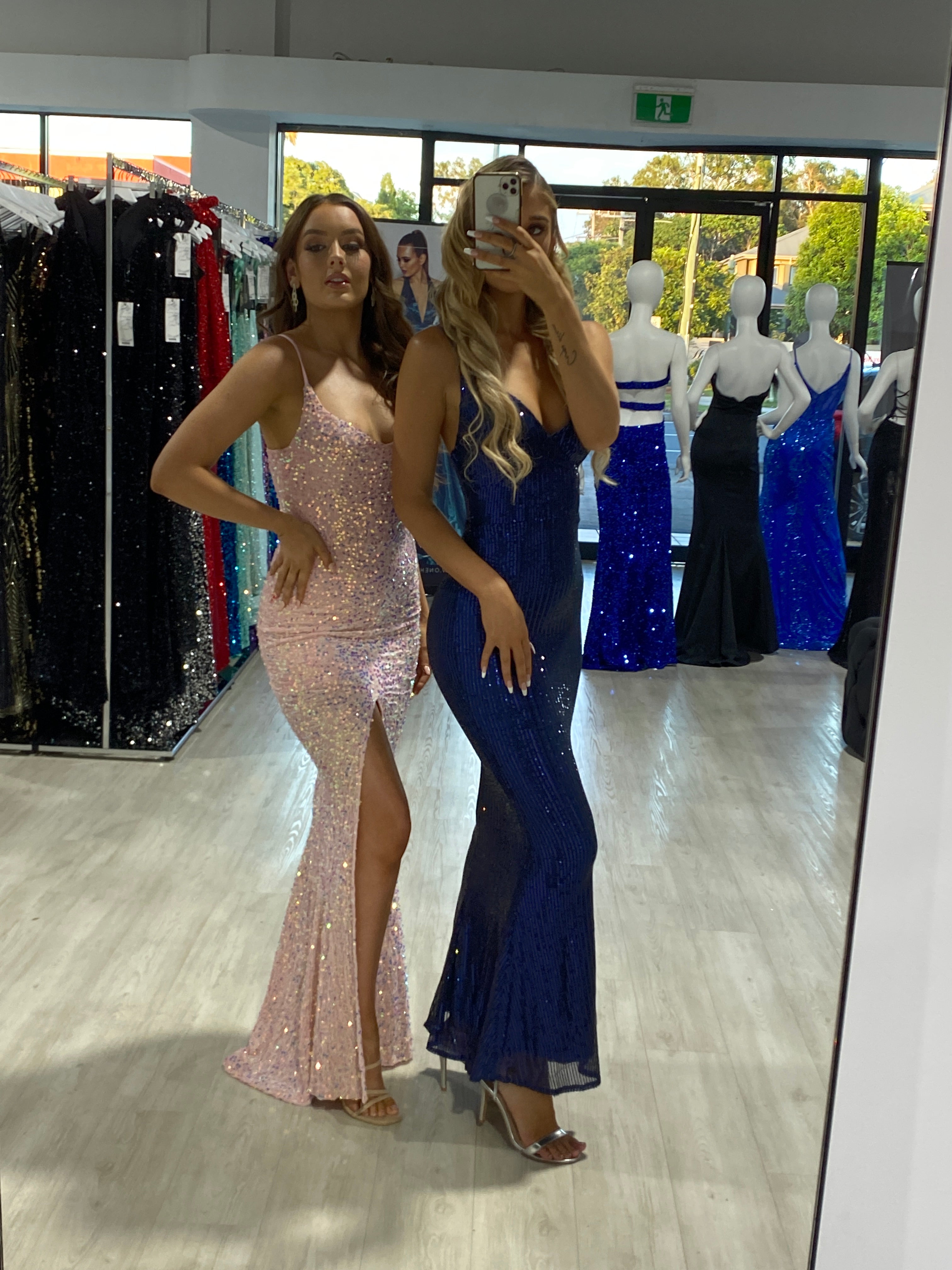 Two women in a boutique trying on sparkly prom dresses, one in navy blue and the other in pink, surrounded by other elegant evening gowns on display.