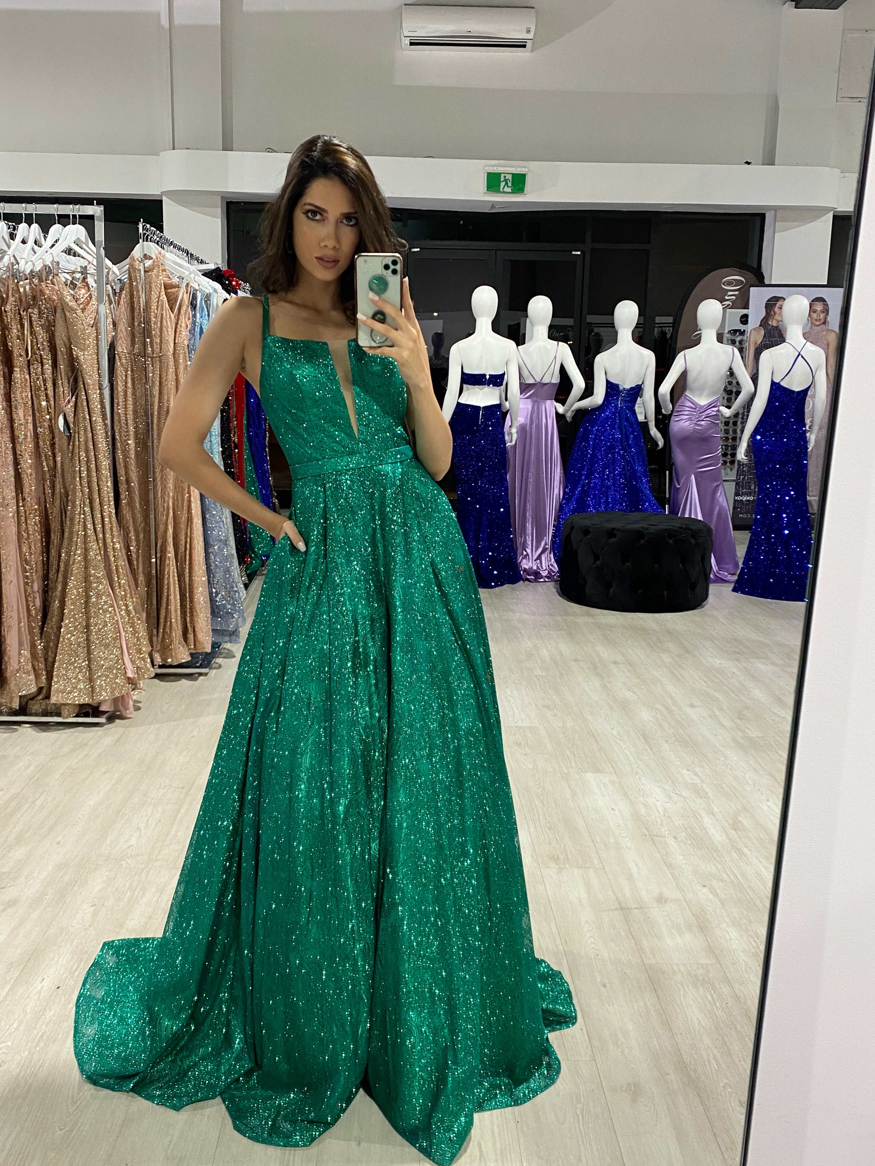 Woman wearing Honey Couture GRETCHEN Green Glitter Ball Gown, an Australian Designer prom dress with pockets and a full skirt, in a dress store.