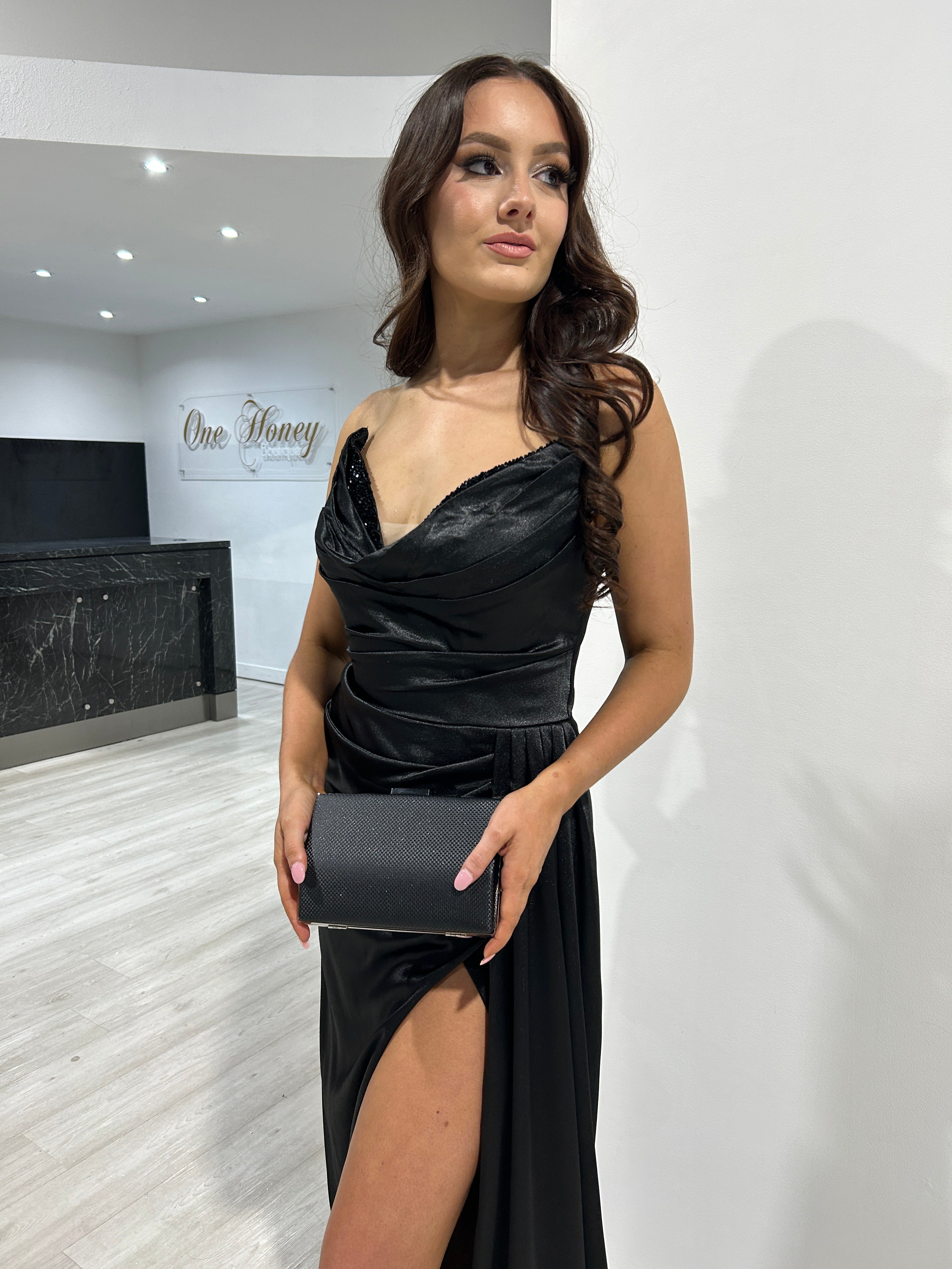 Woman wearing a black satin evening gown with a side slit and cowl neckline, holding a matching clutch. Ideal for formal events.