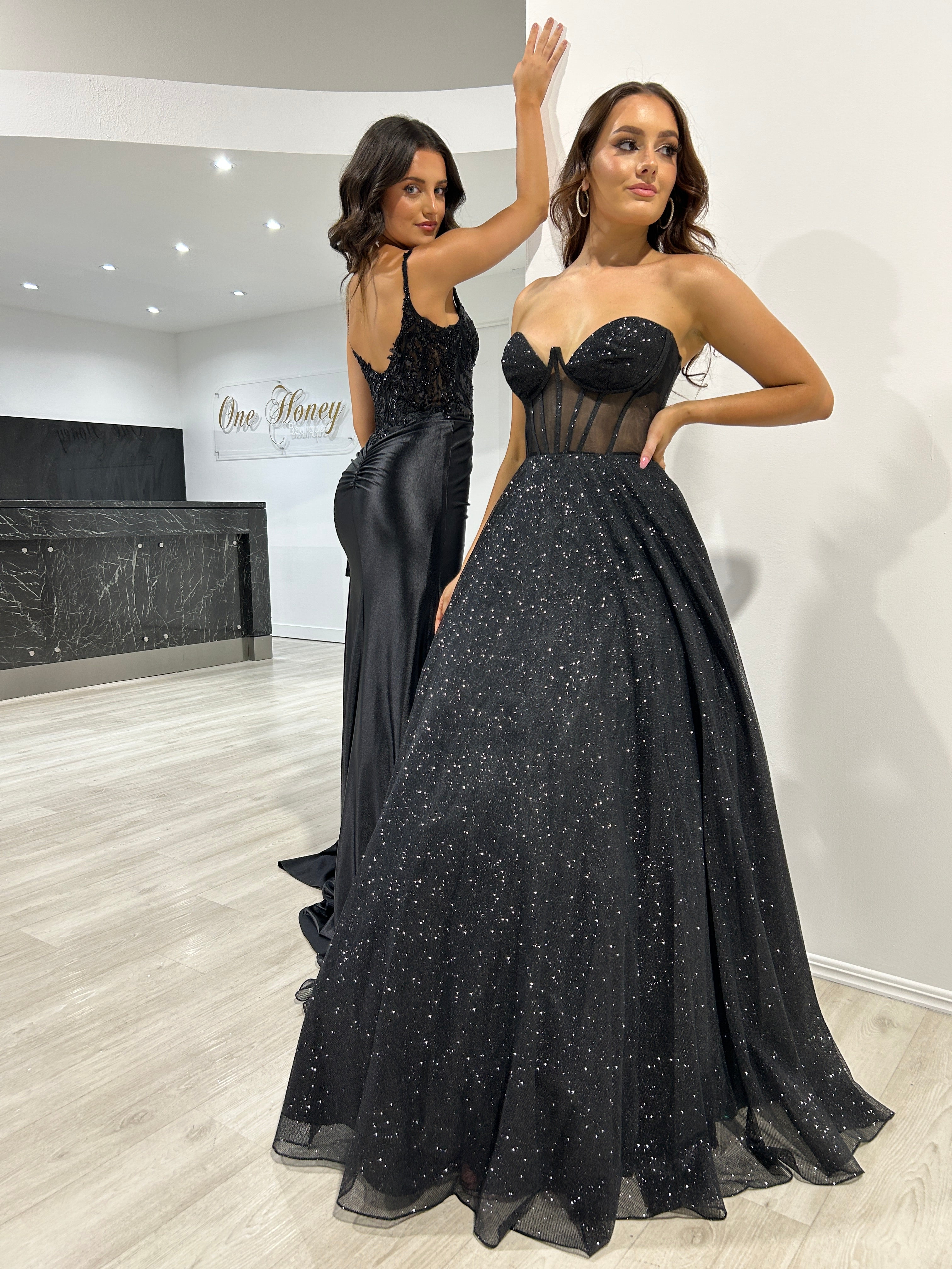 Two women in elegant black evening gowns, one with a glitter corset and tulle skirt, posing in a modern boutique. Brisbane to Gold Coast.