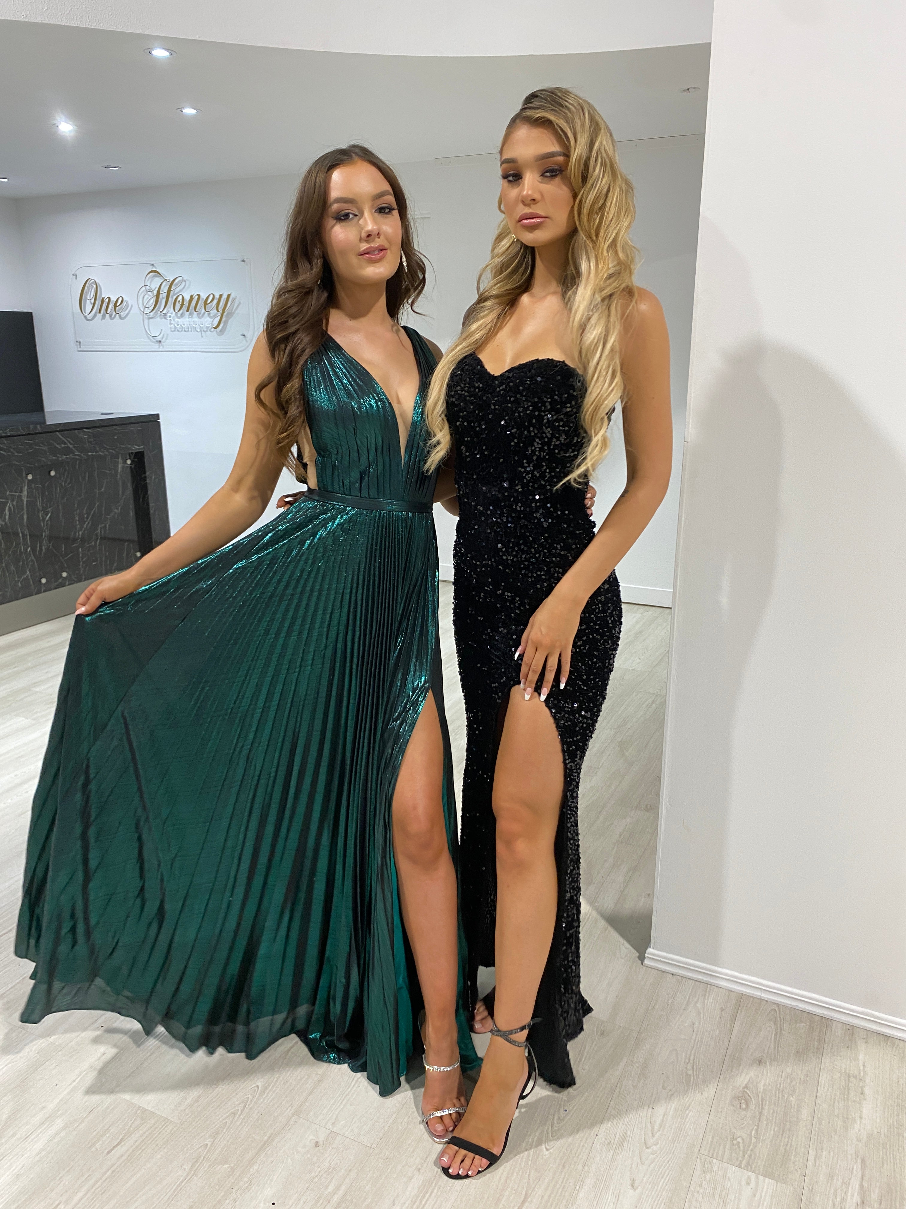 Two women wearing Honey Couture gowns; one in a TRACEY Emerald Green shimmer pleated formal gown, showcasing Australian designer evening wear.