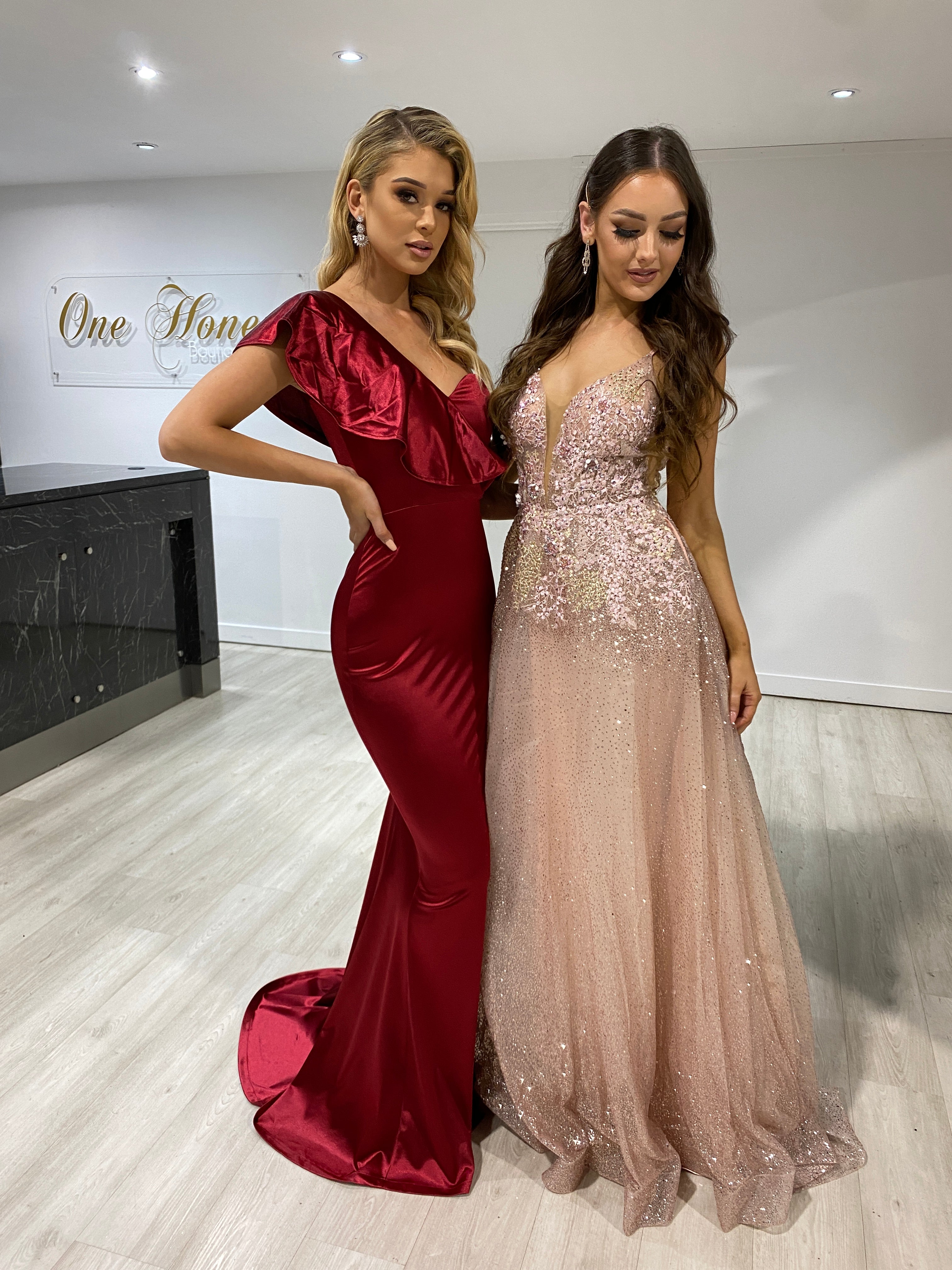 Two women in formal dresses: one in a burgundy one-shoulder Honey Couture mermaid gown, the other in a pink glittering evening dress.
