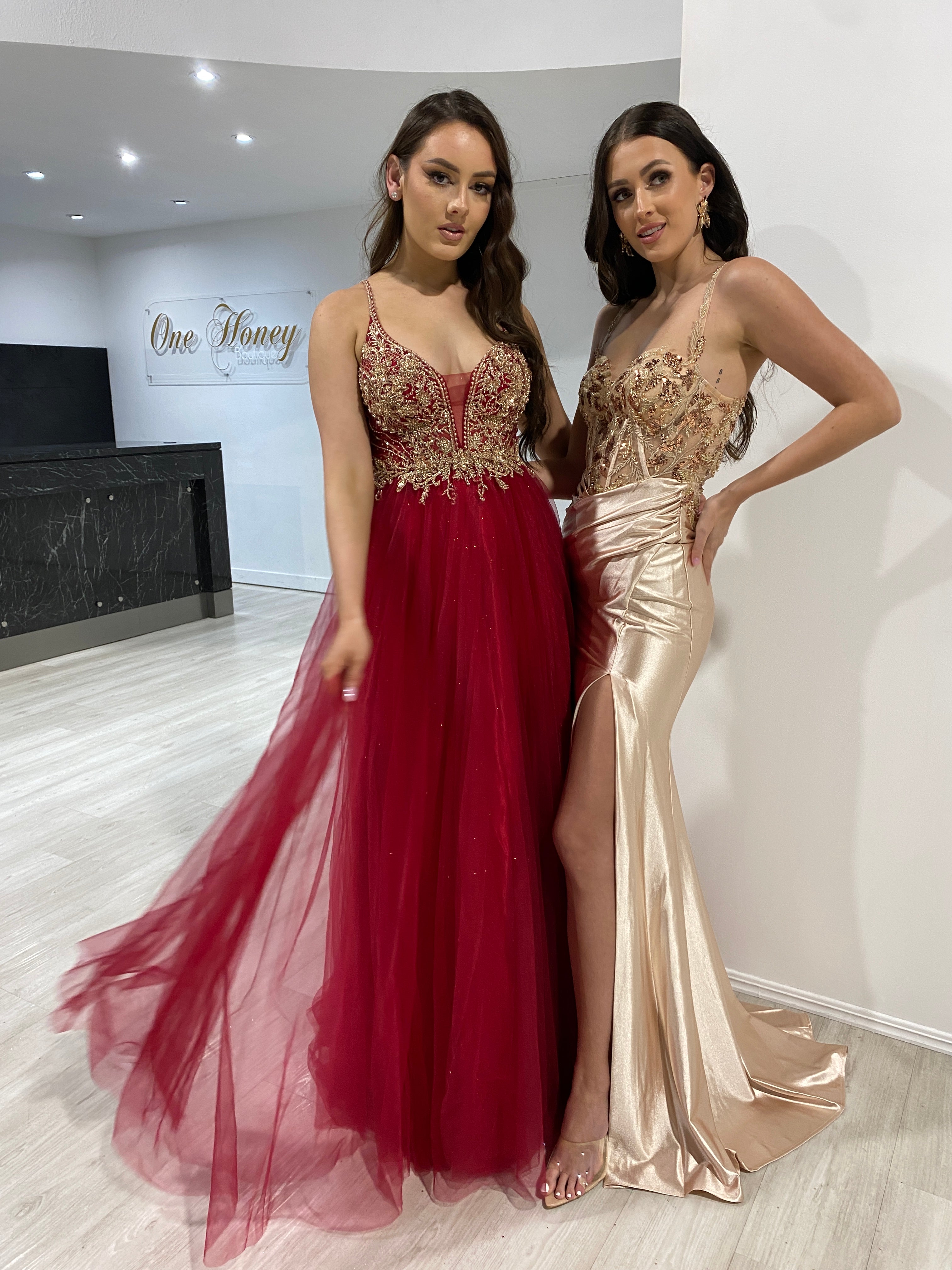 Two women in formal gowns, one in a red beaded tulle dress and the other in a gold embellished satin gown, standing indoors.