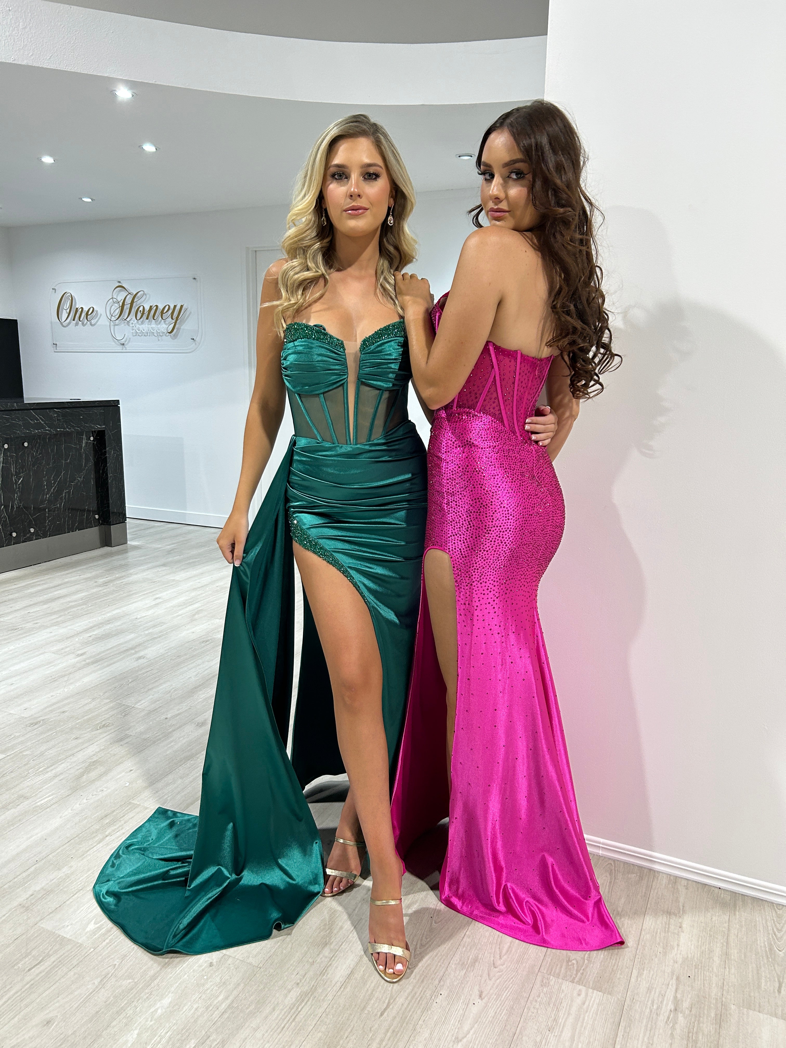 Two women in glamorous strapless evening gowns, one in emerald with sheer boned corset, the other in sparkling pink, on a light wood floor.