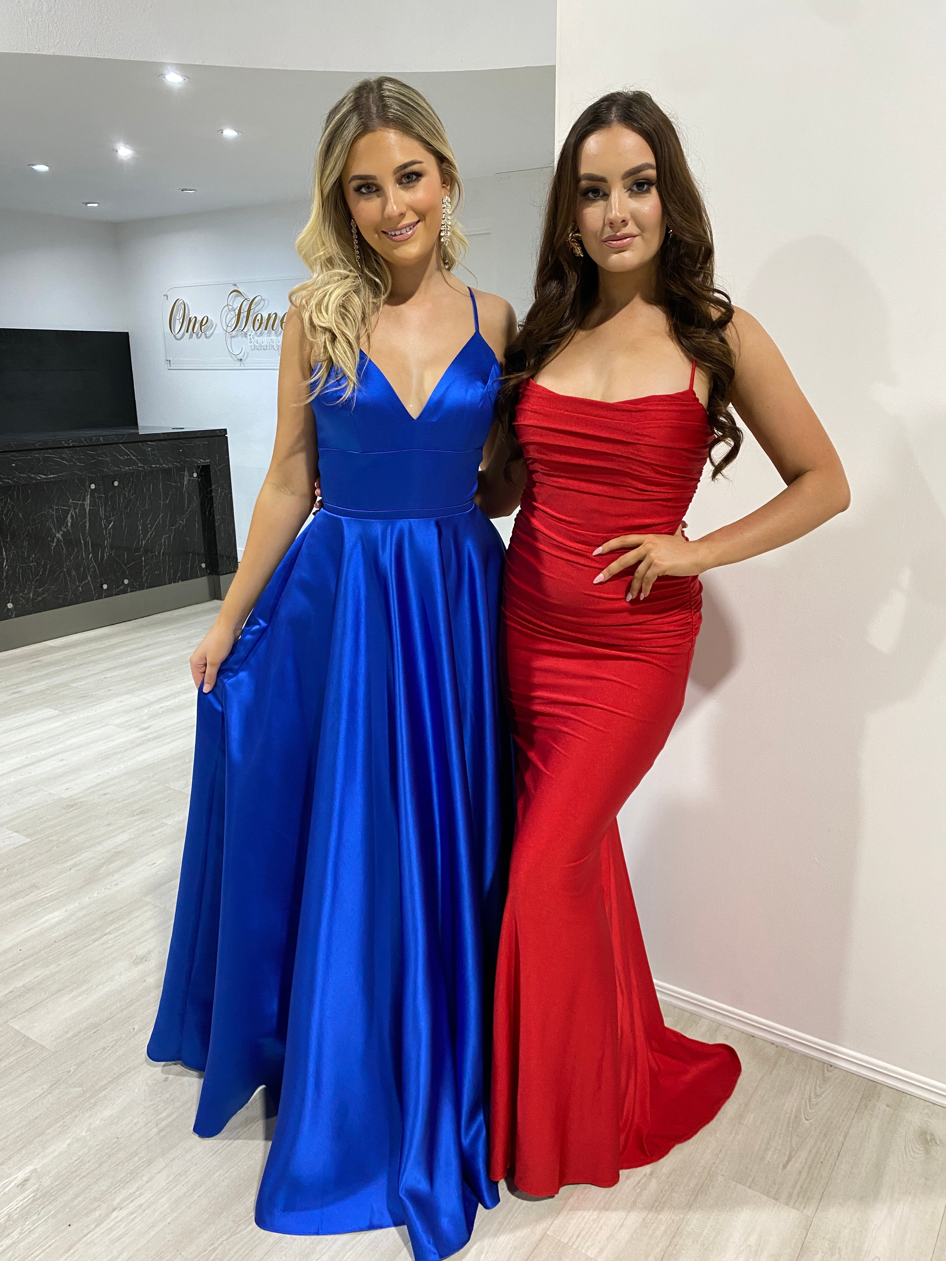 Two women in a formal setting wearing a cobalt blue satin ball gown with a fitted bodice and a red ruched strapless evening gown.