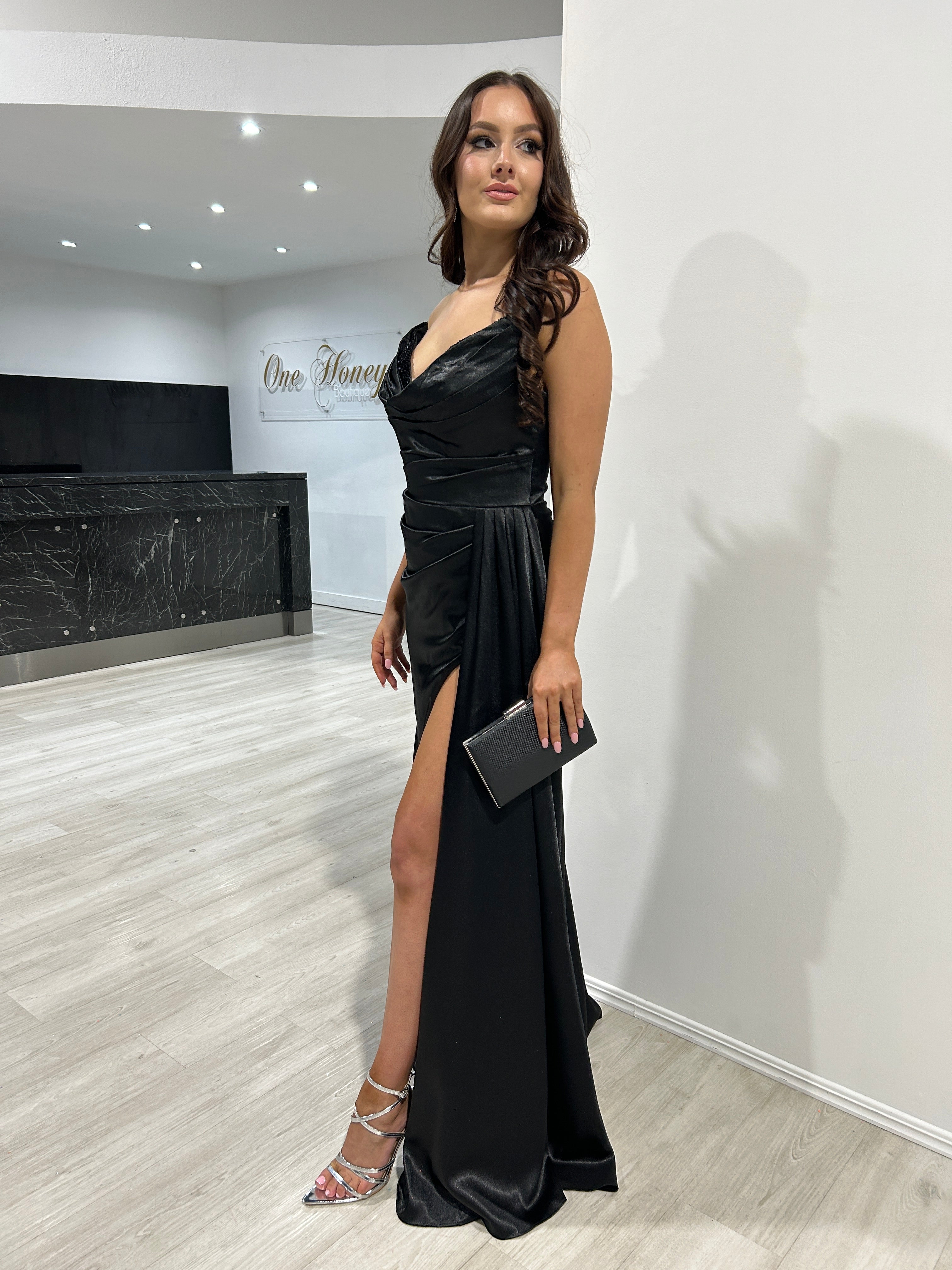 Woman in elegant black evening gown with draped bodice and thigh-high slit, holding a clutch in a chic modern setting.