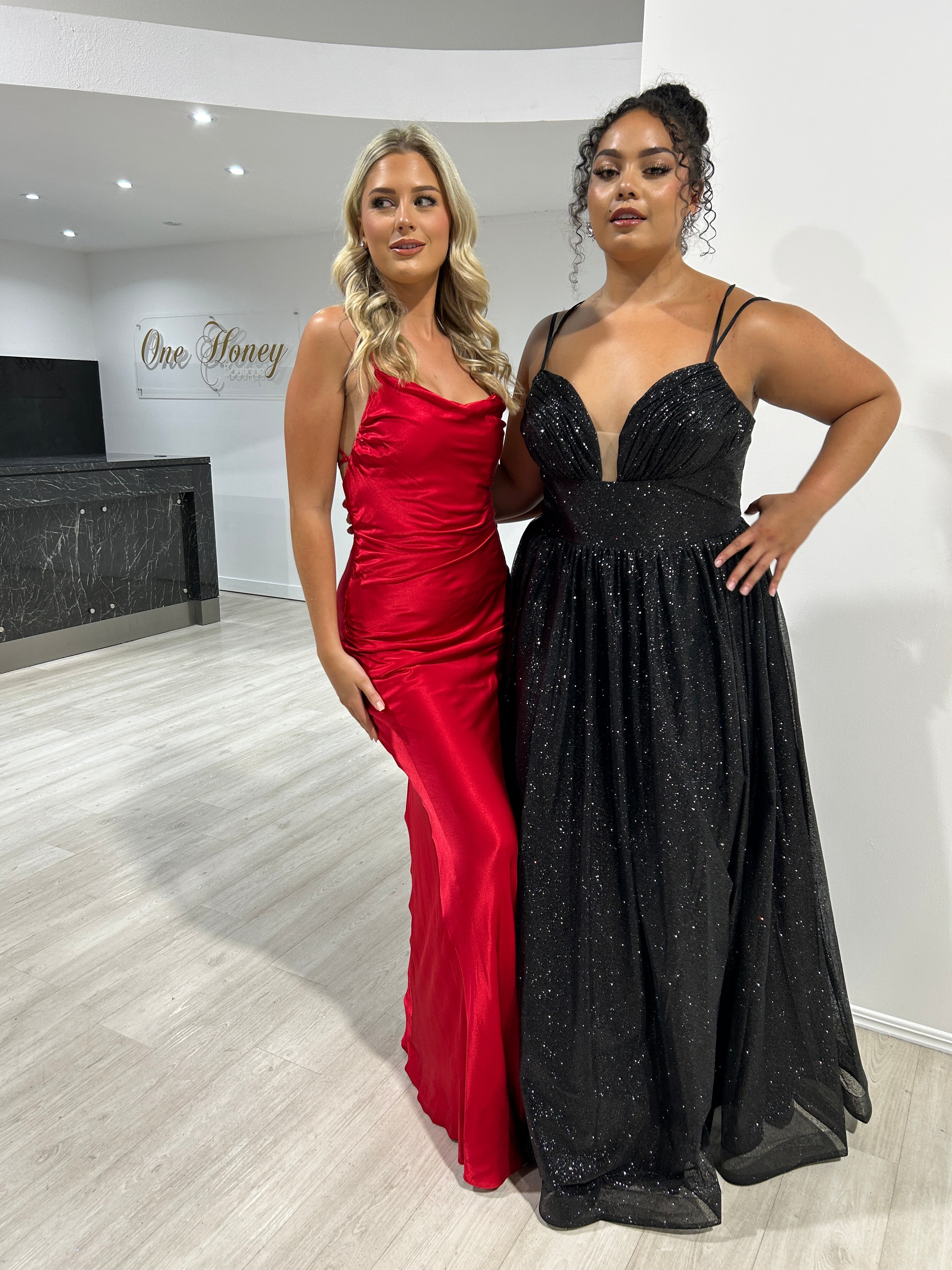 Two women in formal dresses, one wearing a red satin gown and the other in a black glitter ball gown, standing in a modern interior.