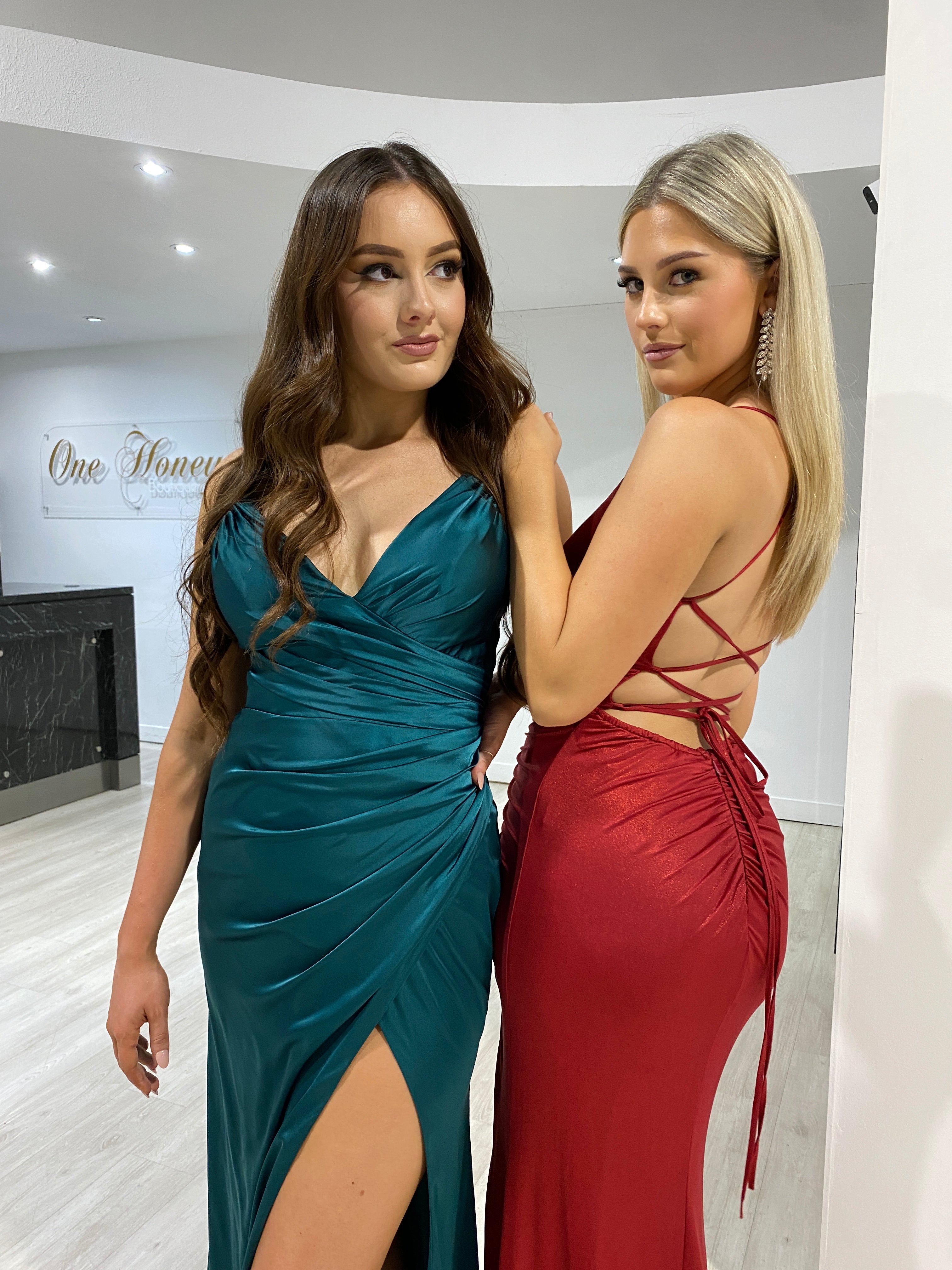 Two models in Honey Couture dresses; one in teal with a leg split, the other in MARI Red Shimmer with lace-up back. Prom and evening gowns.