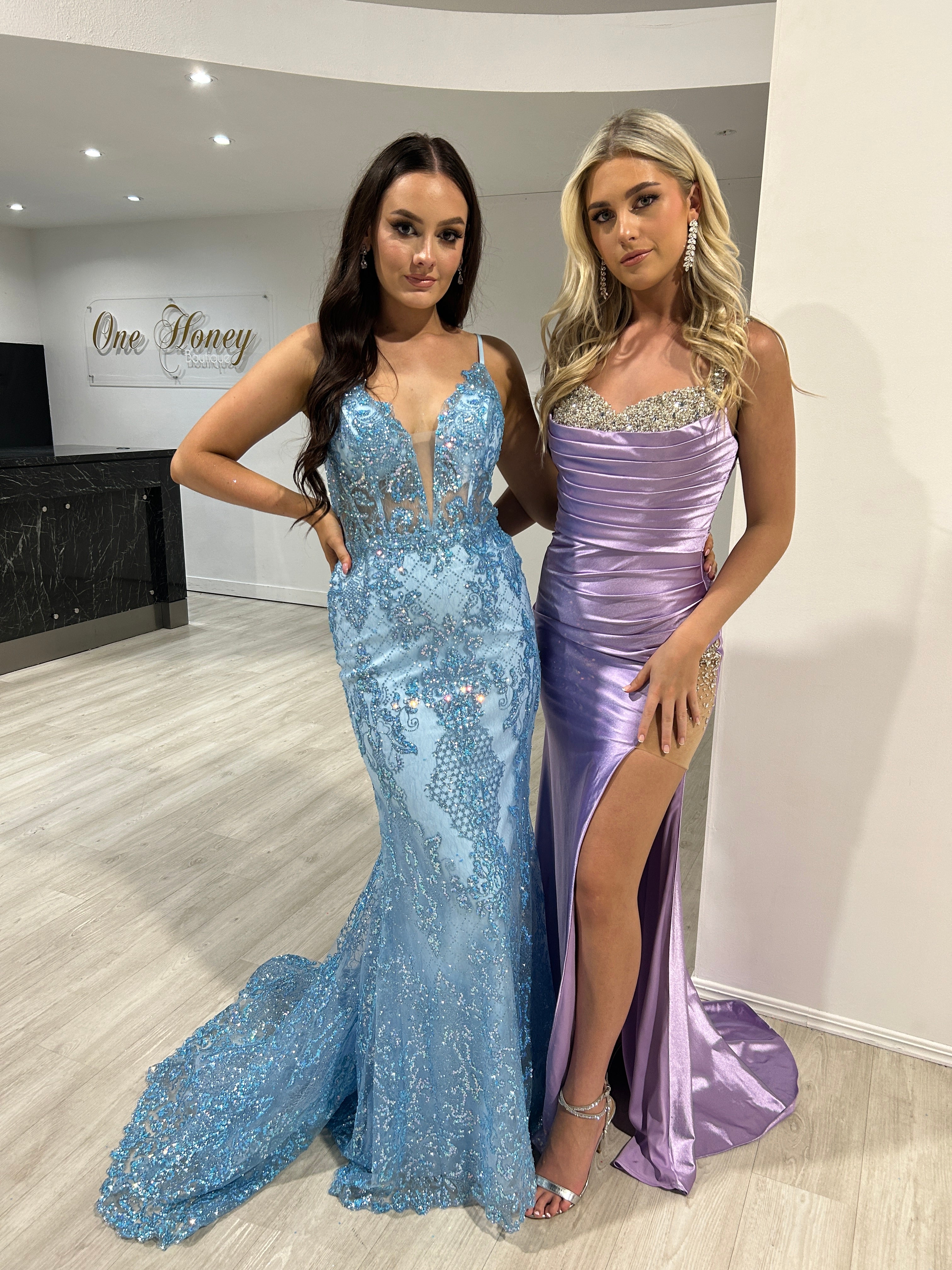 Two women wearing elegant formal gowns, one in a blue lace mermaid dress and the other in a lilac satin gown with a slit, posing indoors.
