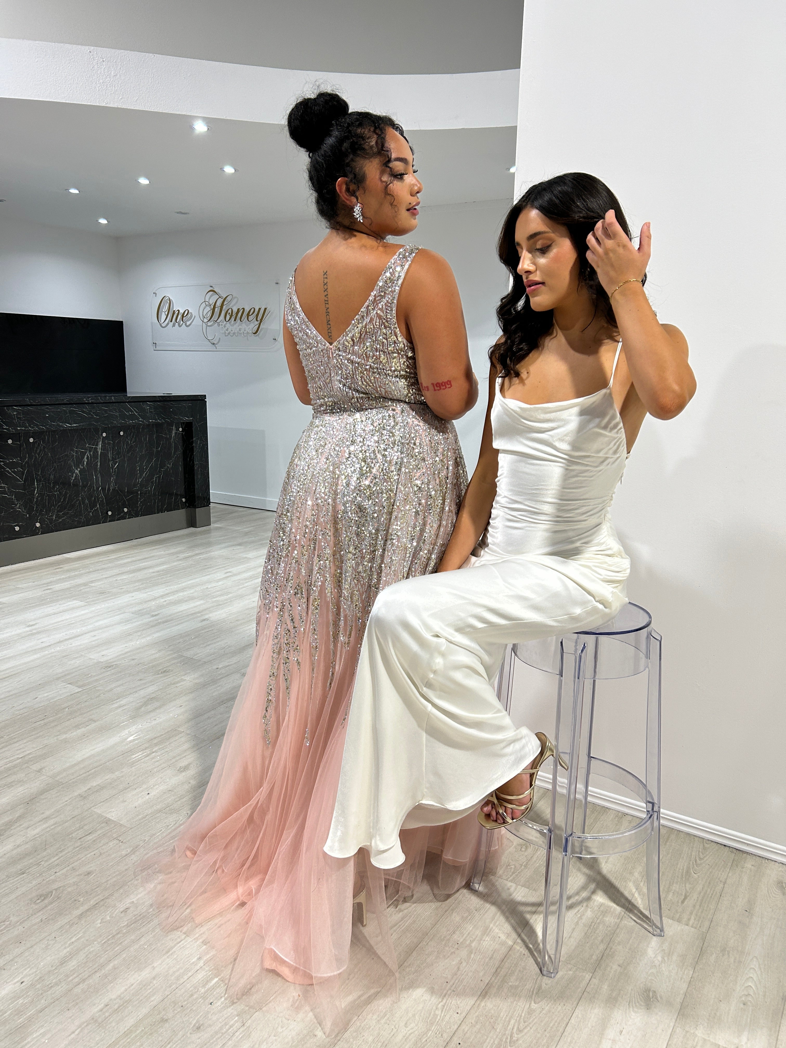 Two elegant women in formal dresses, one in a shimmering silver and pink gown, the other in a sleek white satin gown, in a stylish setting.