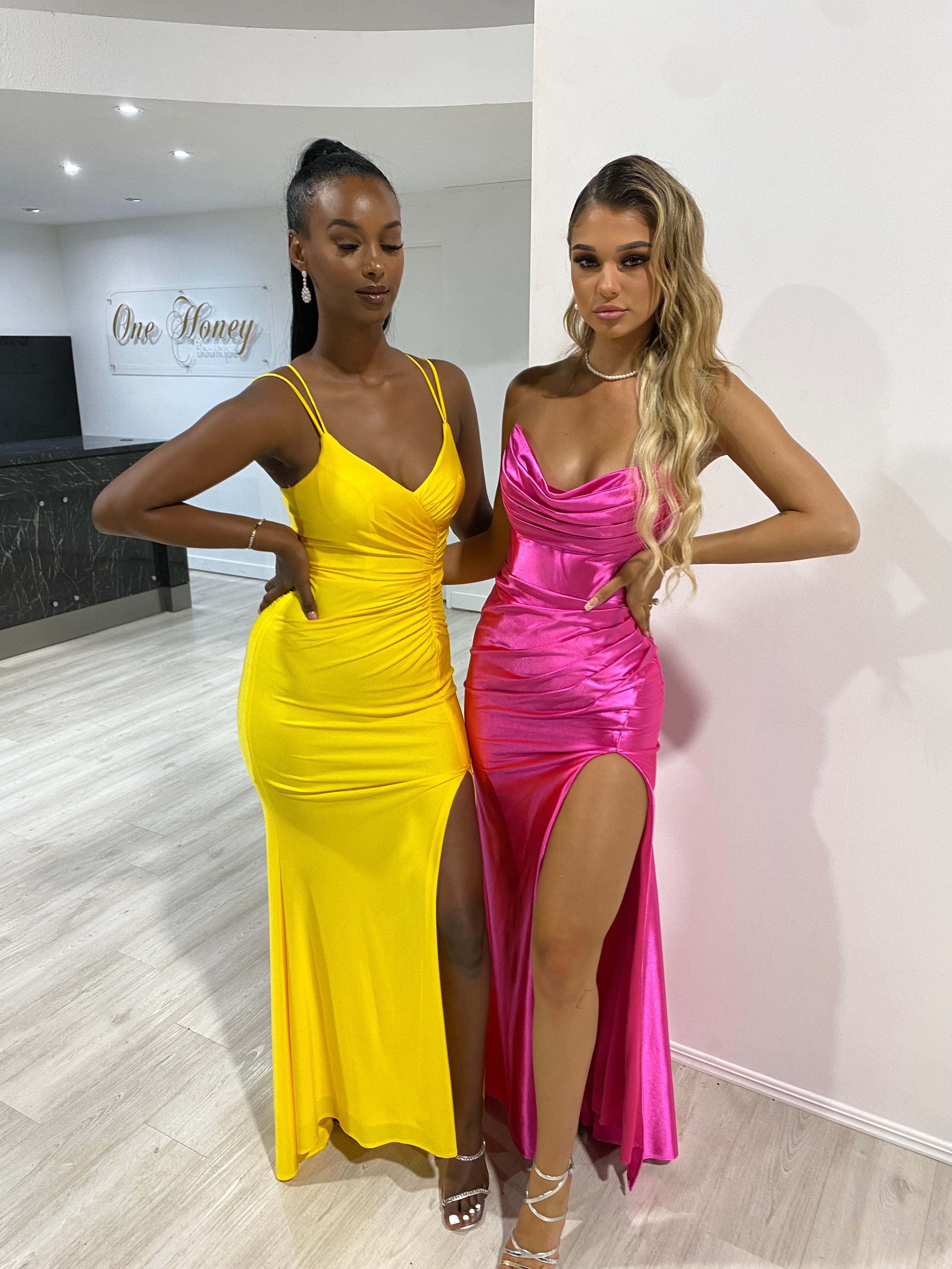 Two women wearing vibrant Honey Couture formal gowns, one in neon yellow and the other in hot pink, perfect for prom or evening events.