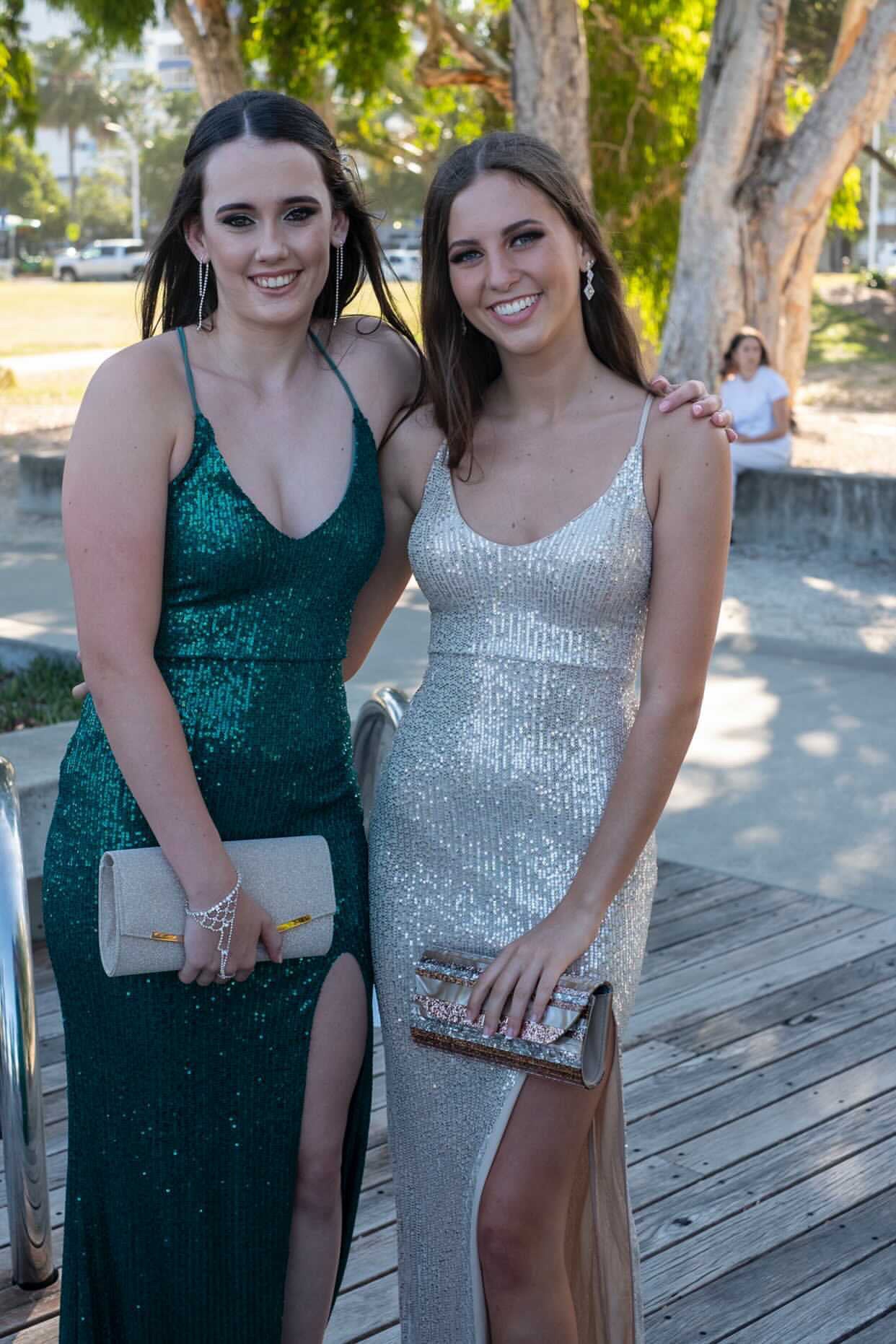 Two women in sequin gowns, one in emerald green and the other in silver, standing outdoors during the day, holding clutch purses.