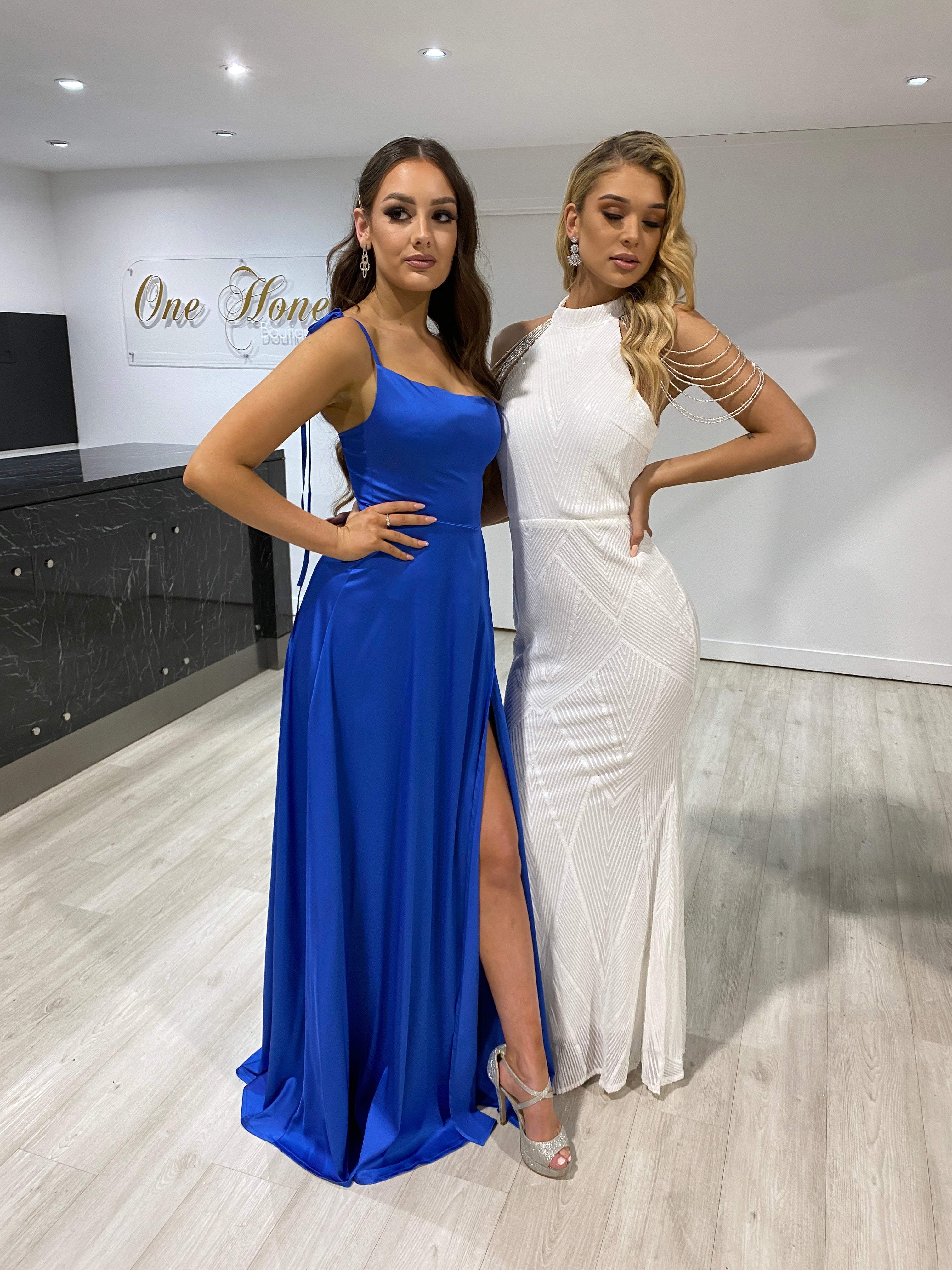 Two models wearing a blue XENIA Honey Couture evening gown and a white formal dress, showcasing affordable Australian designer prom dresses.