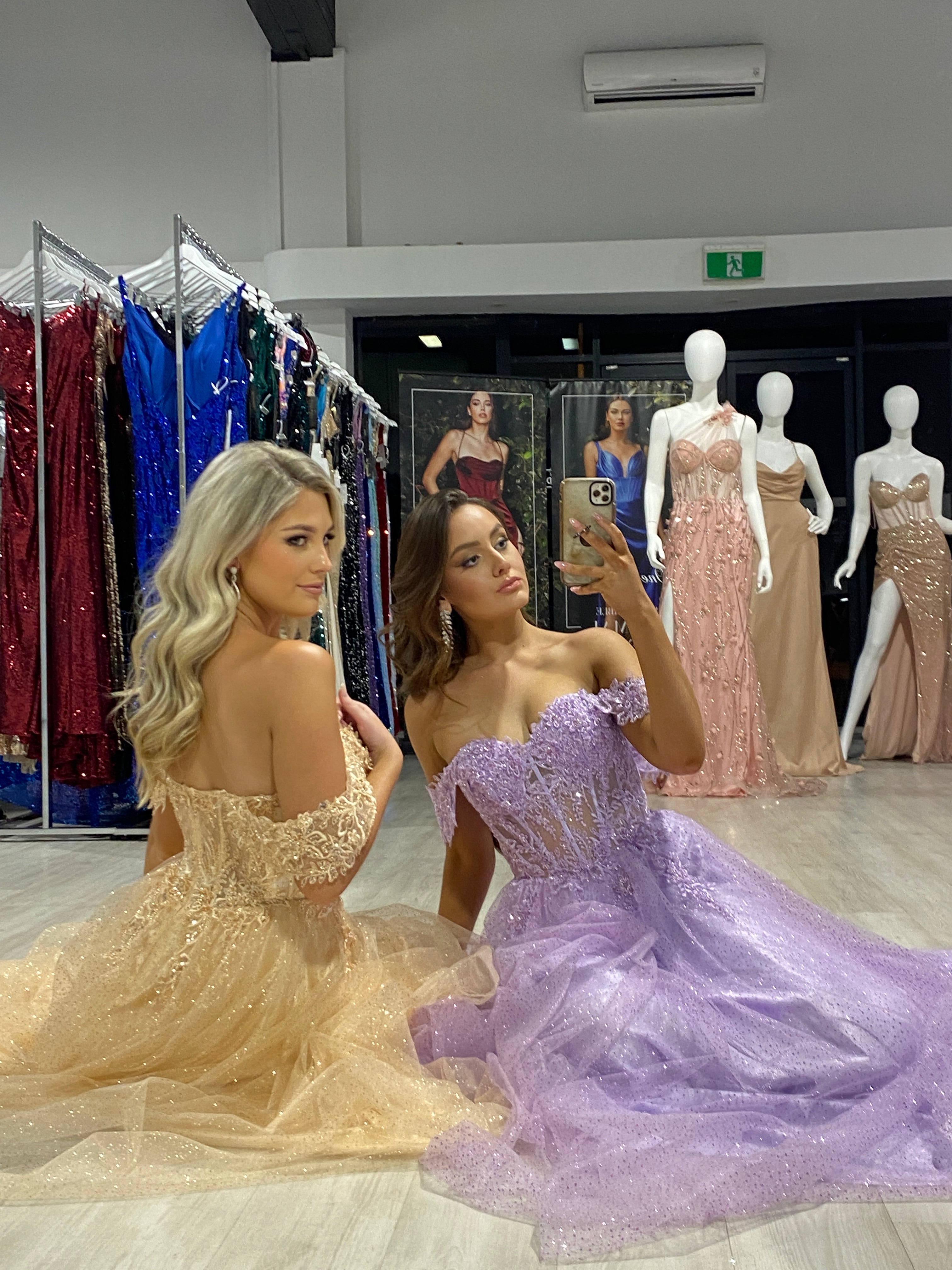 Two women in glittery off-the-shoulder dresses, one in gold and one in lavender, sit on the floor of a formal dress store.