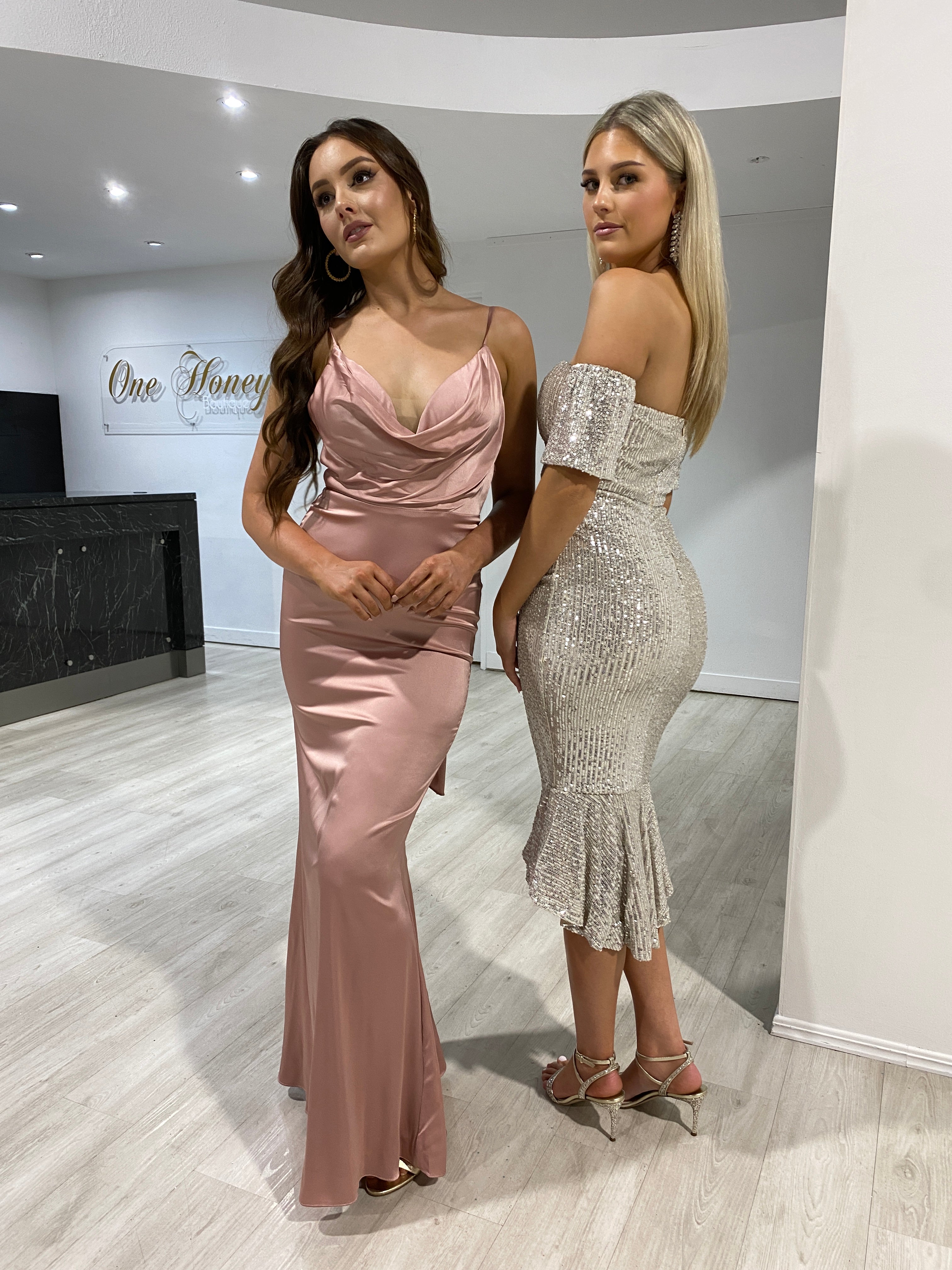 Two women wearing Honey Couture dresses: one in a pink evening gown and another in the NEW YORK Platinum Sequin Off Shoulder Midi Dress from Australian Designer.