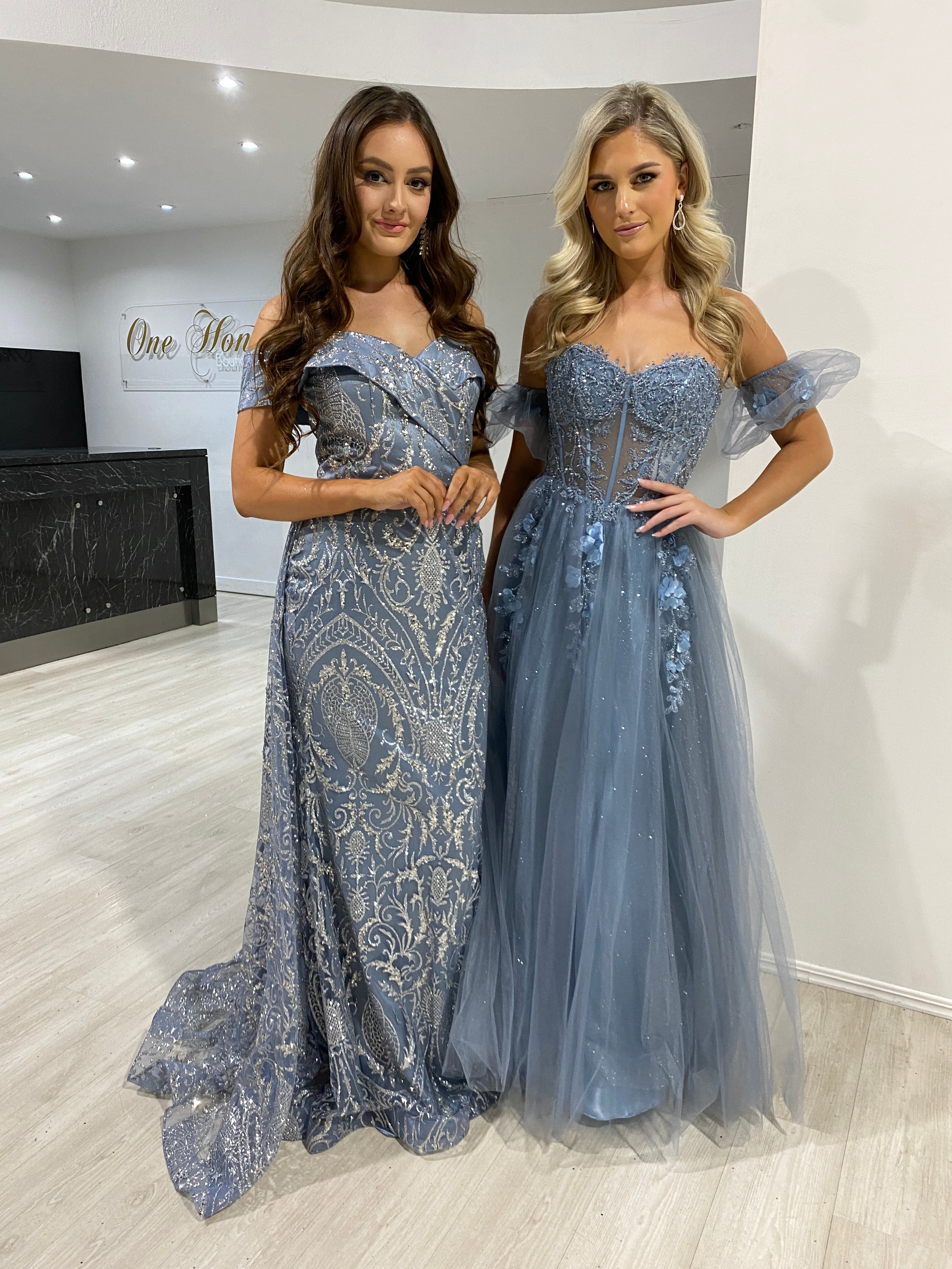 Two women modeling Honey Couture TALITA Smokey Blue Off the Shoulder Shimmer Ballgowns, designed by Australian designers. Perfect for prom or evening events.