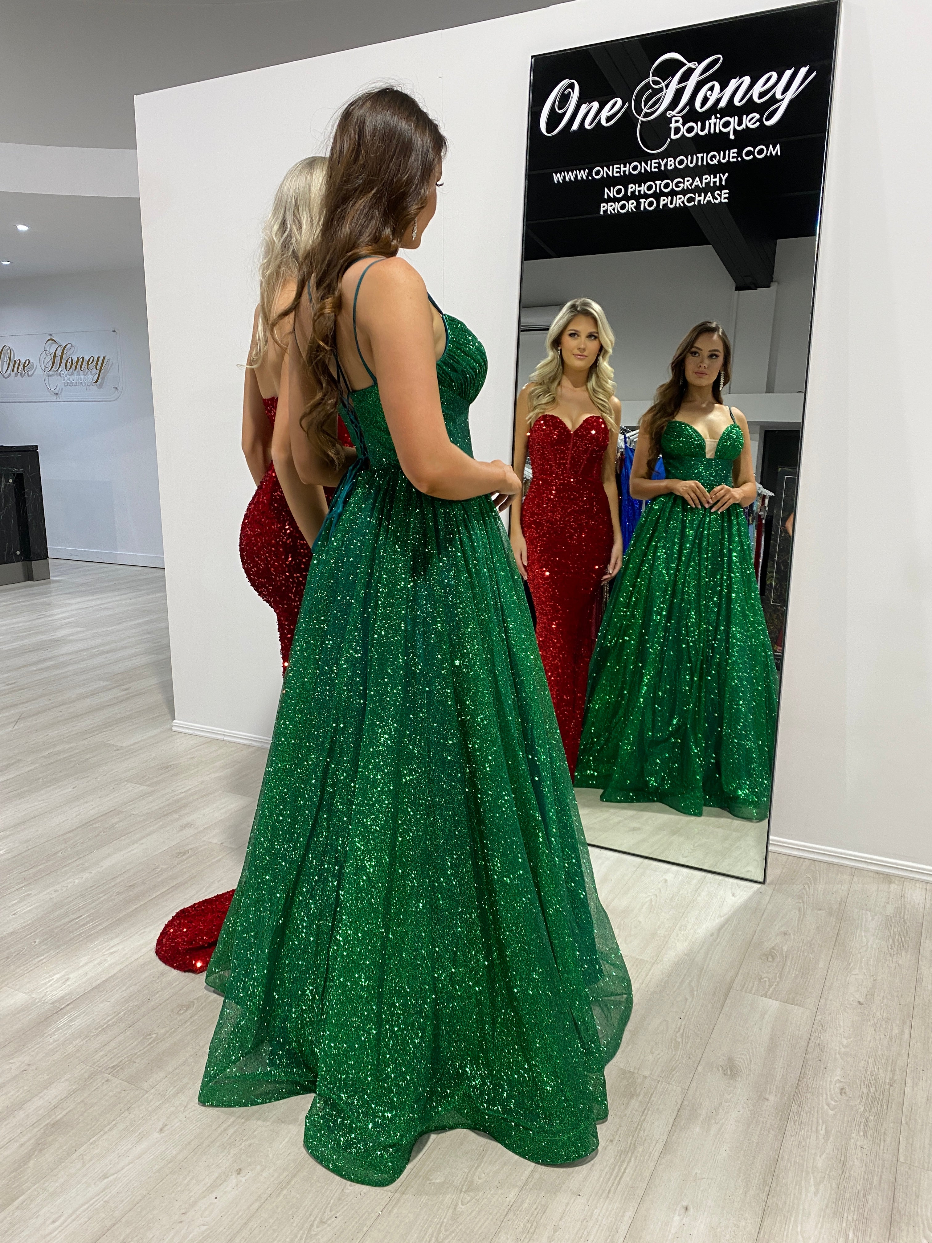 Women wearing Honey Couture evening gowns in front of a mirror, featuring the MADDISON red strapless mermaid dress and a green sequin gown.