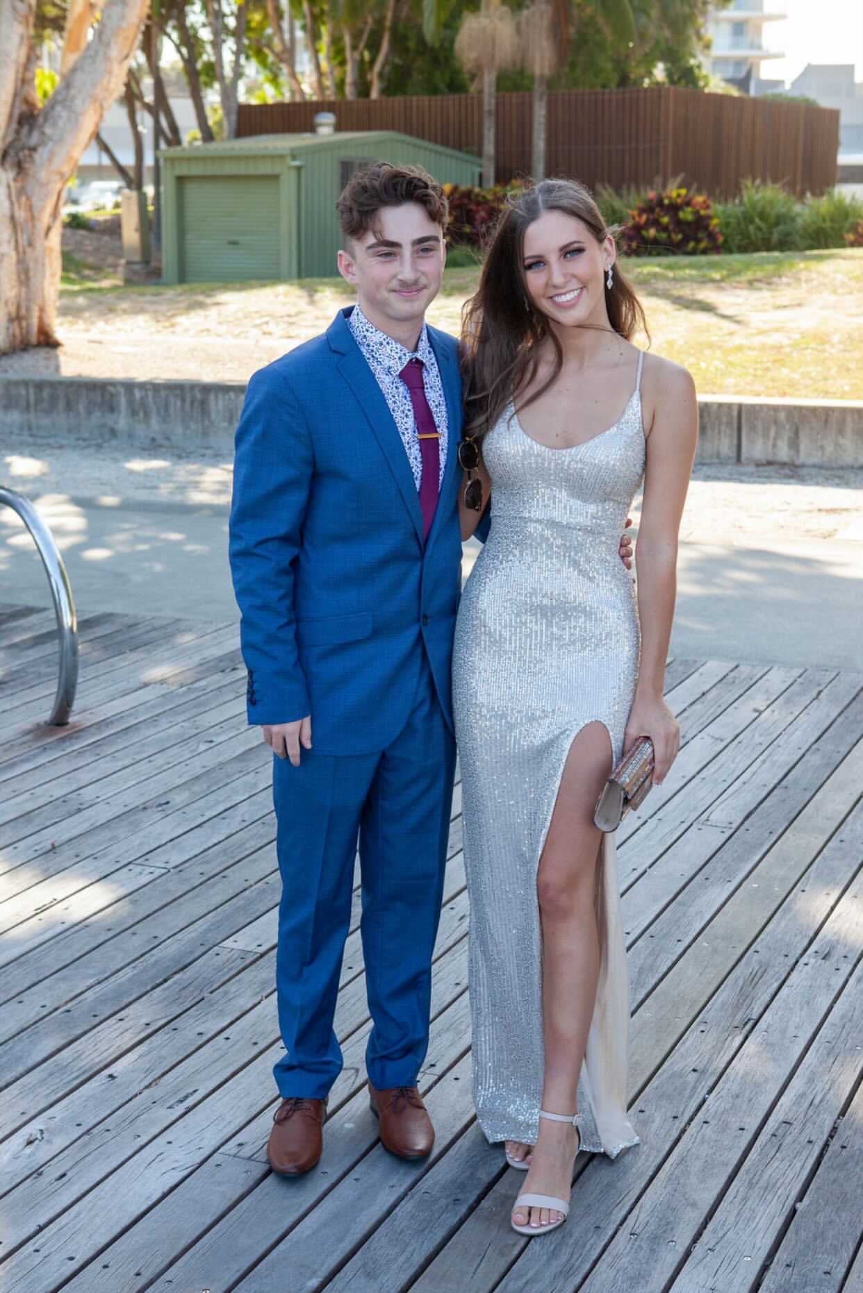 Woman in a silver sequin gown with lace-up back standing next to man in a blue suit outdoors on a boardwalk.