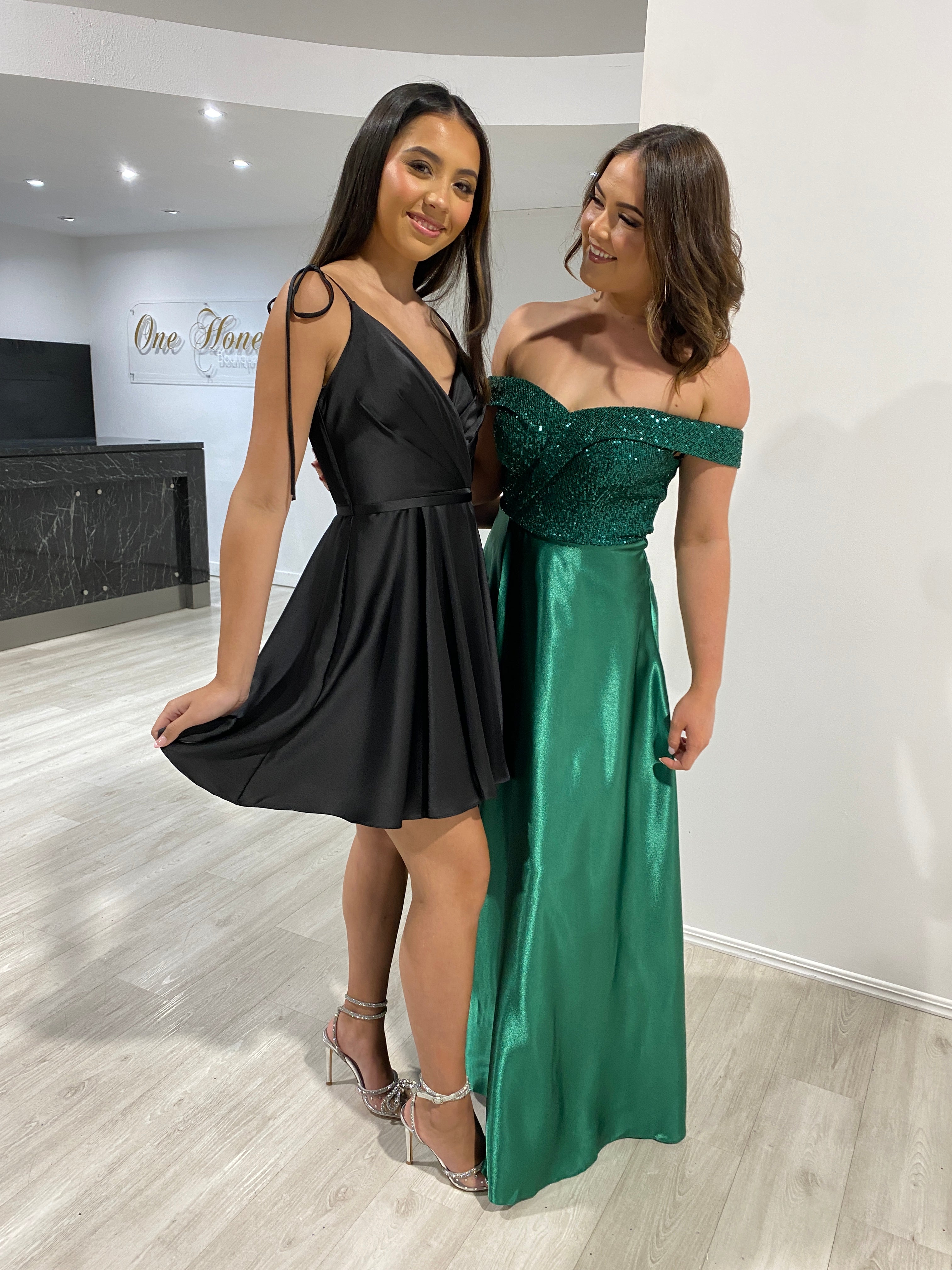 Two women wearing formal dresses; left in Honey Couture ESTELLA Black Silky Circle Skirt Mini Party Dress, right in green evening gown.