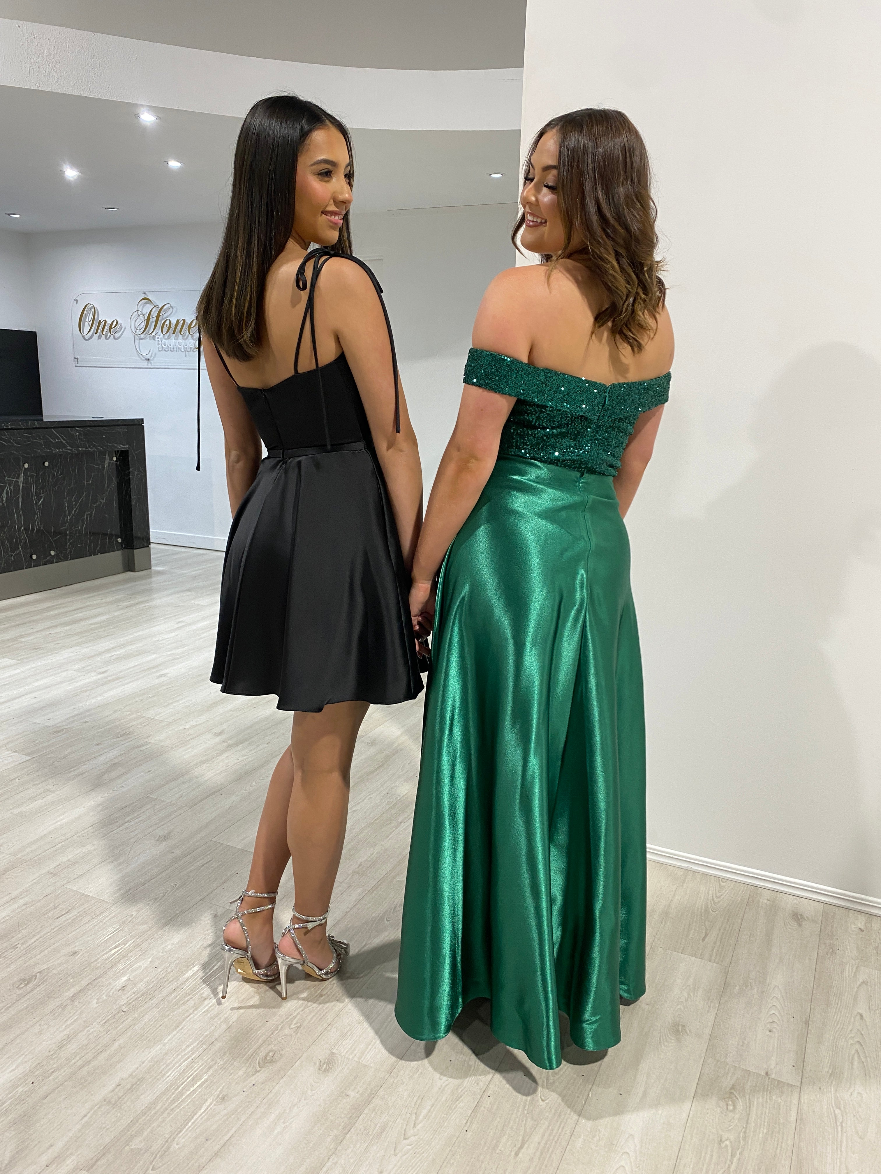Two women wearing party dresses, one in black mini with bow straps and one in green evening gown, perfect for prom or events, from Australian Designer Honey Couture.