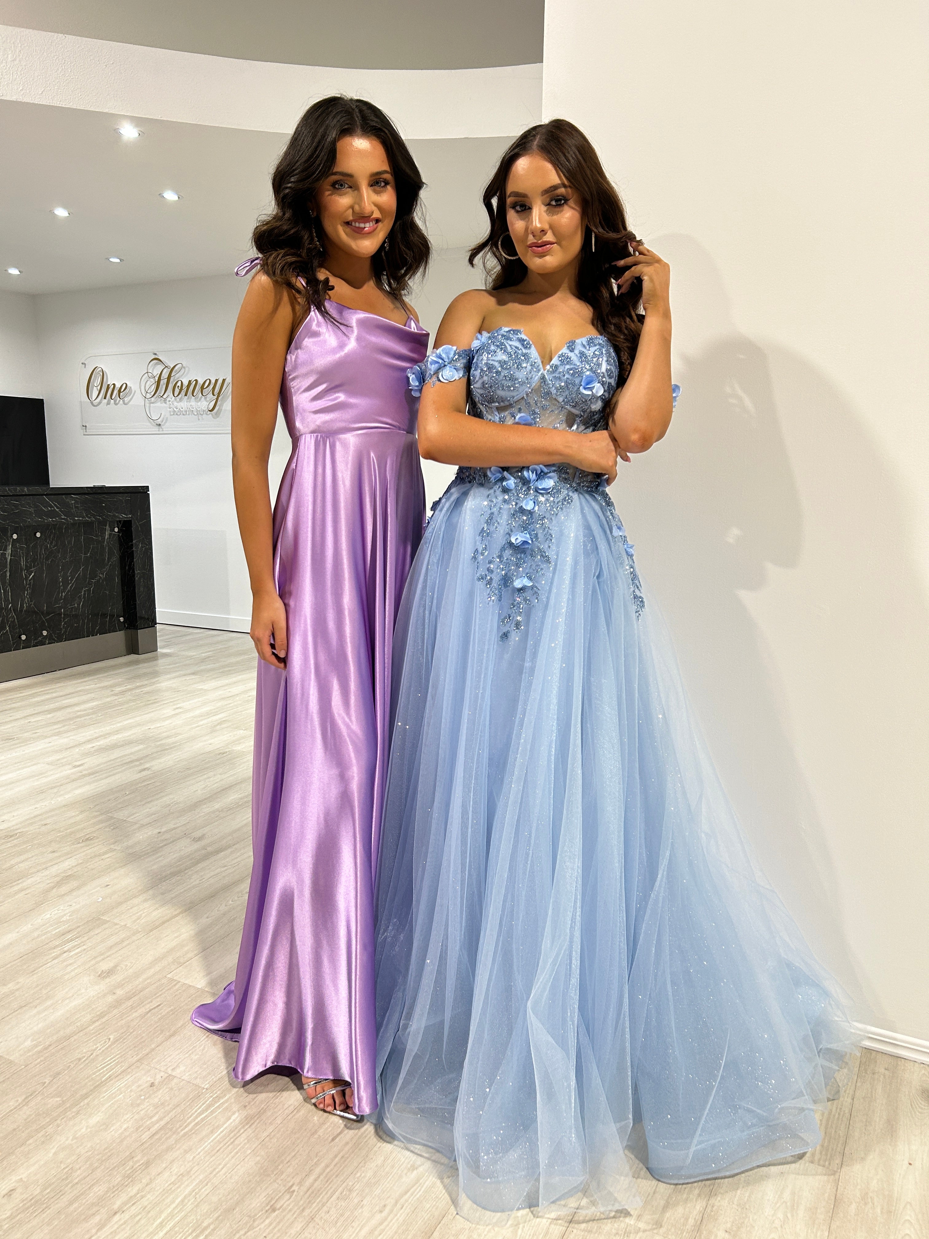 Two women in formal gowns, one in a purple satin dress and the other in a blue off-shoulder glitter A-line dress by Honey Couture.