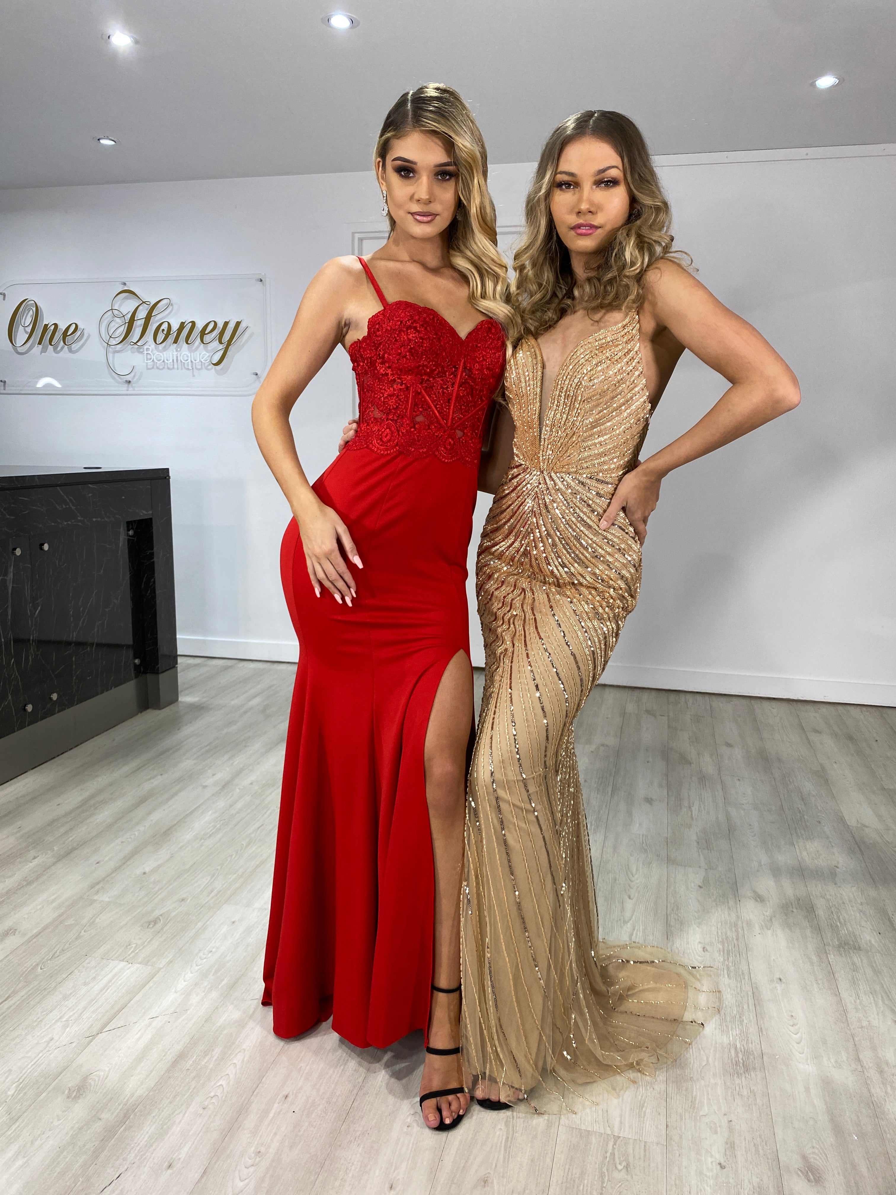 Two women wearing elegant formal dresses, one in a red lace gown with a slit and the other in a gold embellished gown, posing in a showroom.
