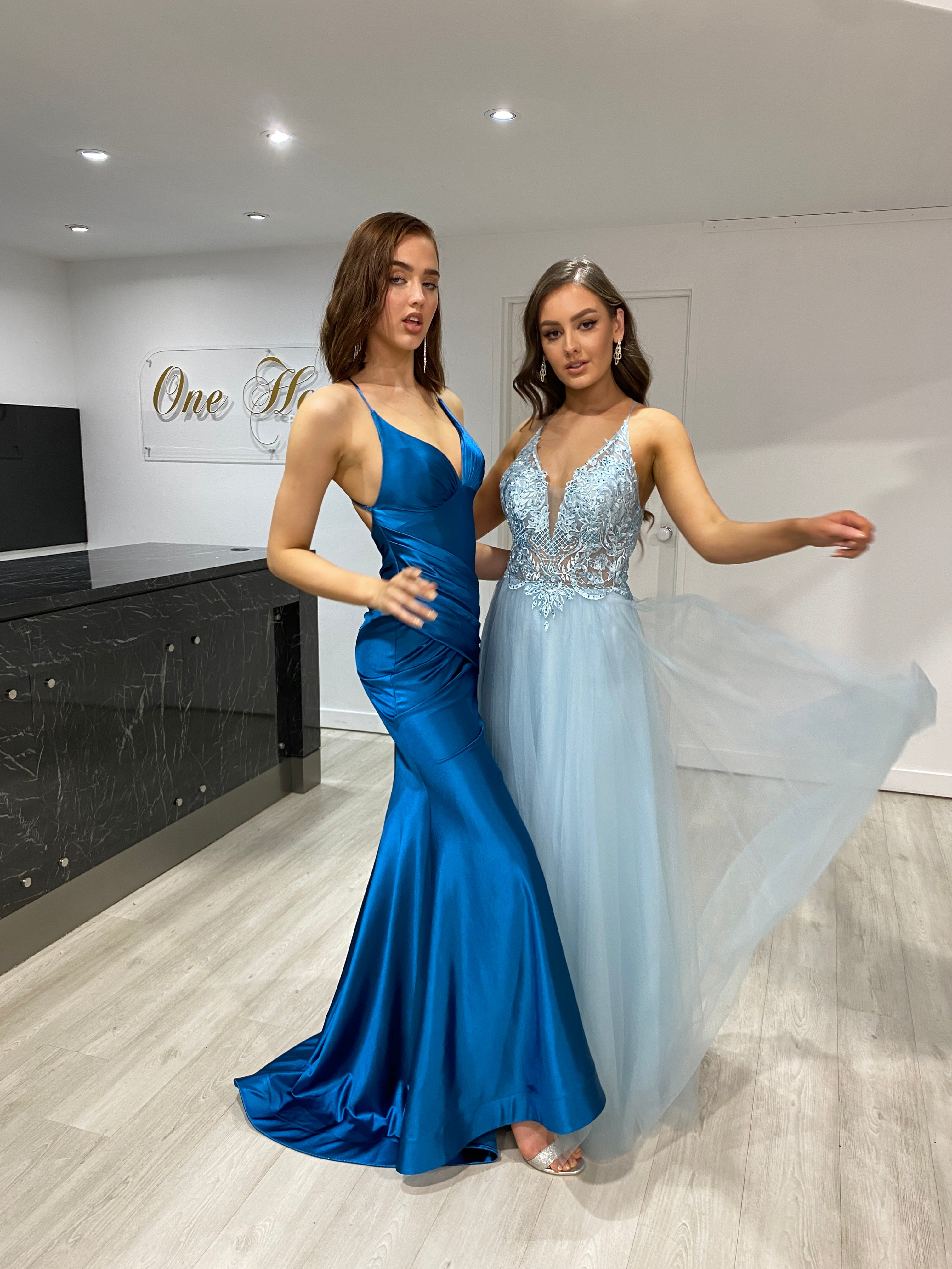 Two women in elegant formal gowns; one in a royal blue satin dress, the other in a Honey Couture INEZ baby blue tulle lace gown.