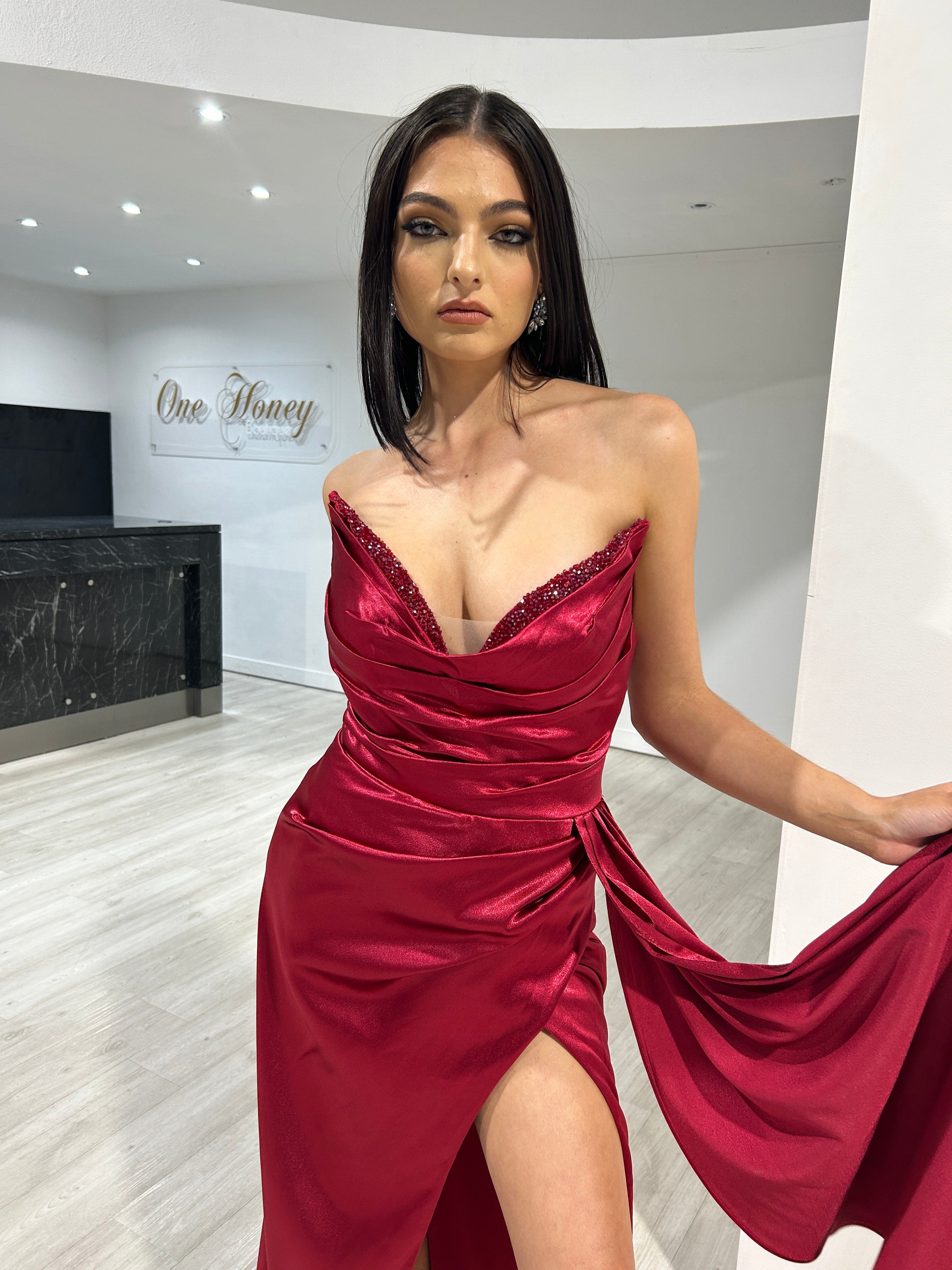 Woman in elegant red satin evening gown with draped design and deep neckline posing indoors at One Honey Boutique.
