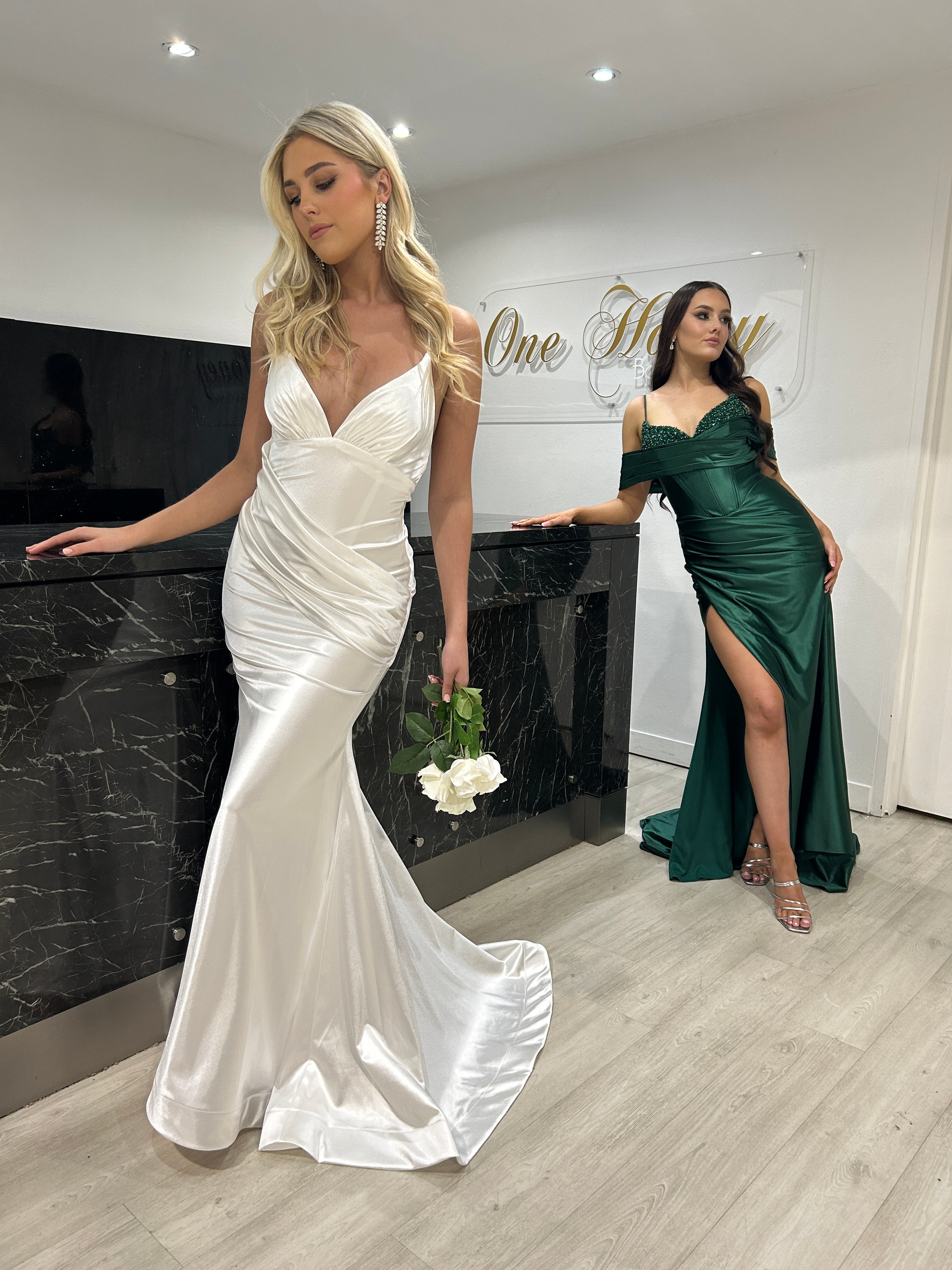 Elegant women in satin formal dresses, white gown with ruched bodice and green dress with thigh-high slit, standing by a marble counter.
