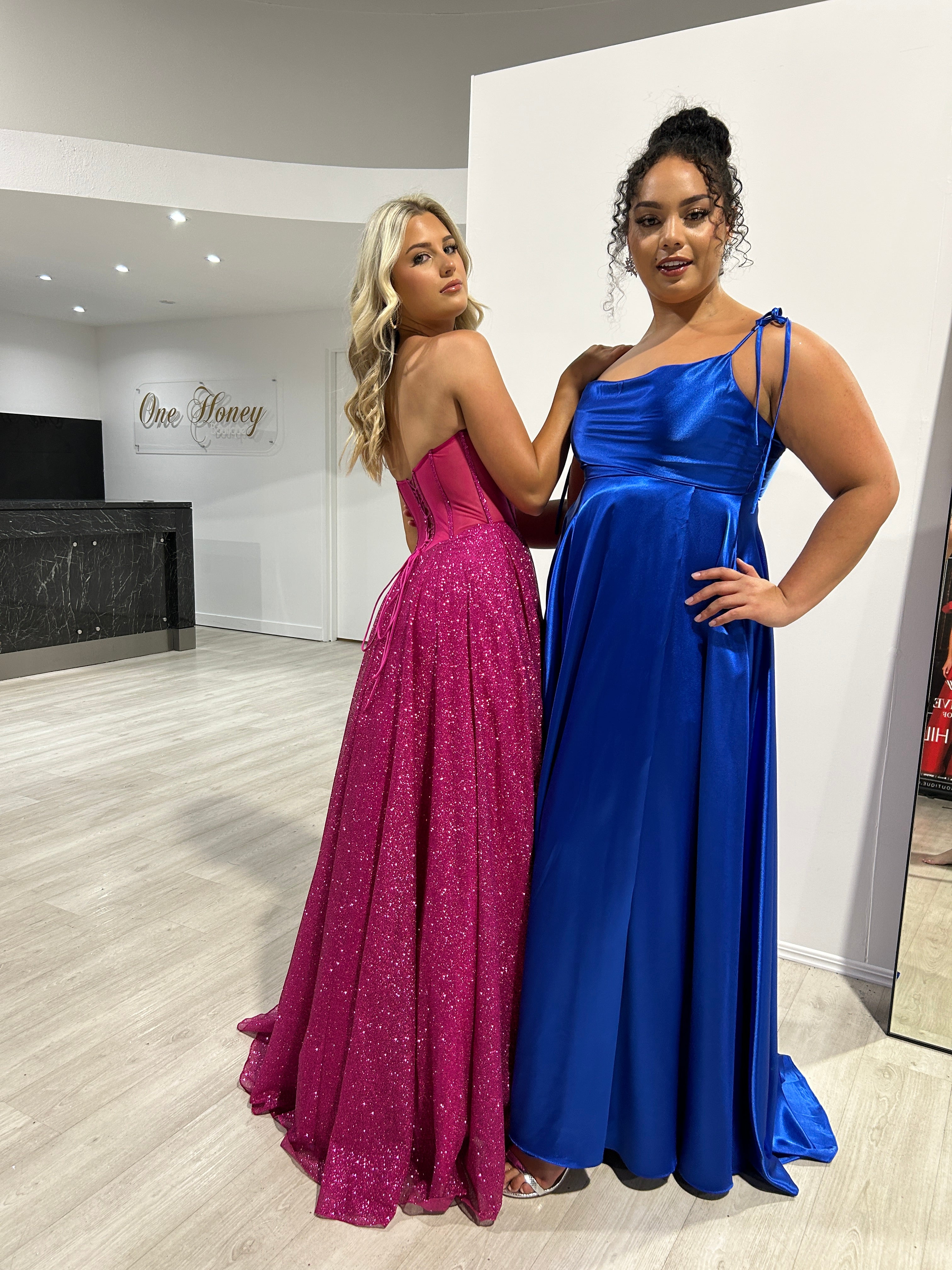 Two women in vibrant formal gowns, one in a purple glitter strapless dress, the other in a blue satin tie-strap gown. Fashion showroom display.