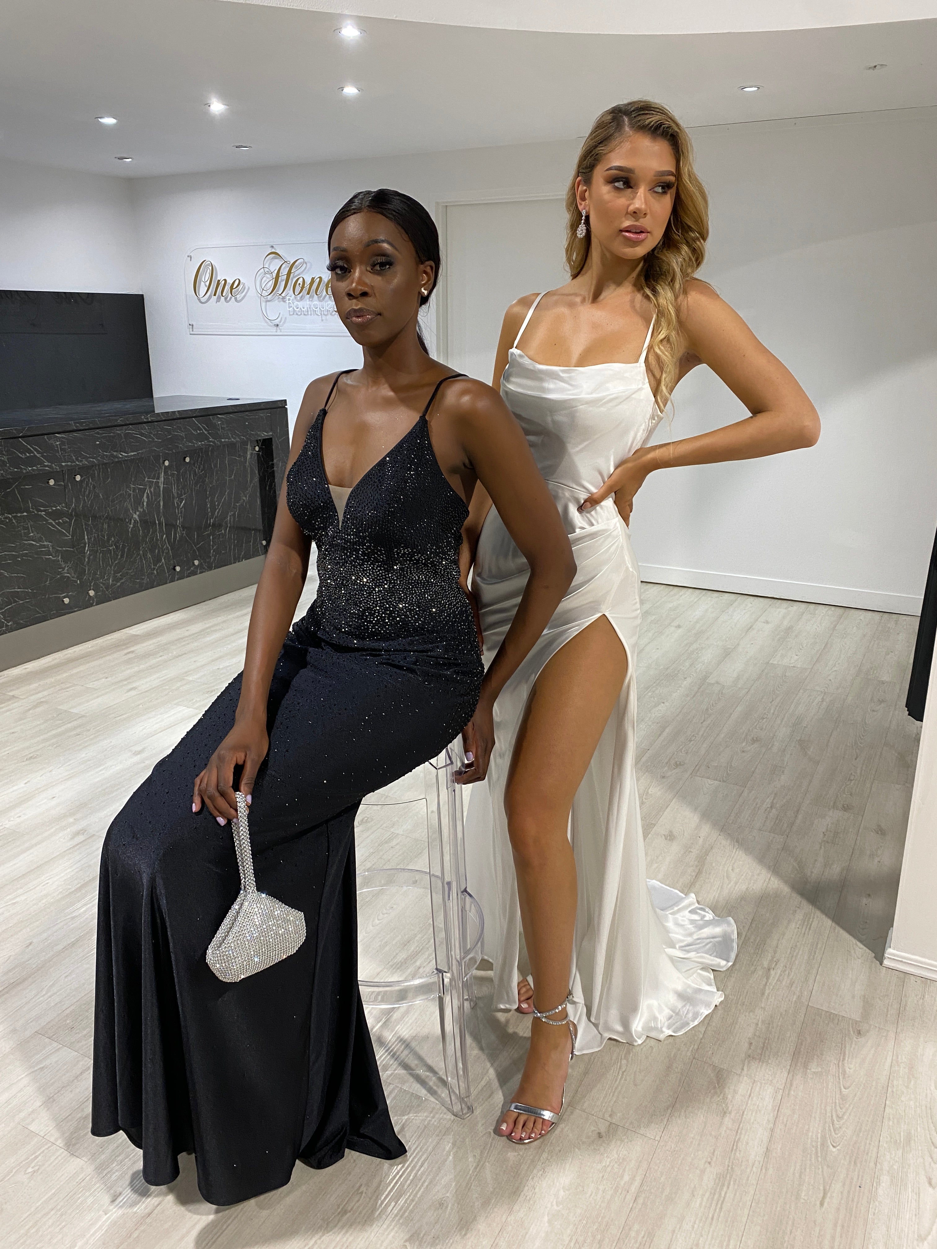 Two women in elegant evening gowns, one in black and one in Honey Couture QUINN off white satin lace-up dress, showcasing prom and evening fashion.
