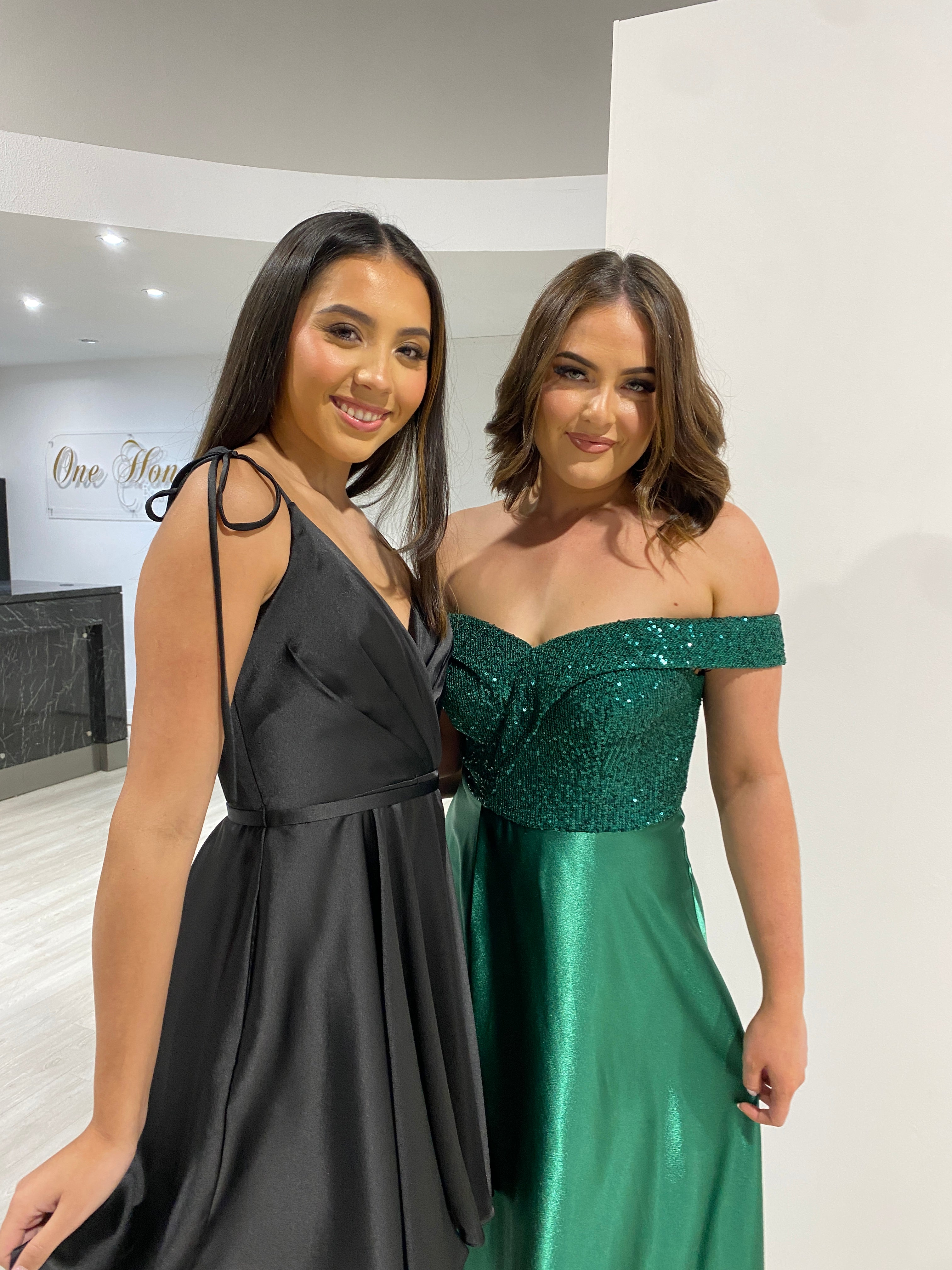 Two women modeling the ESTELLA Black Silky Circle Skirt Mini Party Dress by Honey Couture and a green off-shoulder gown.
