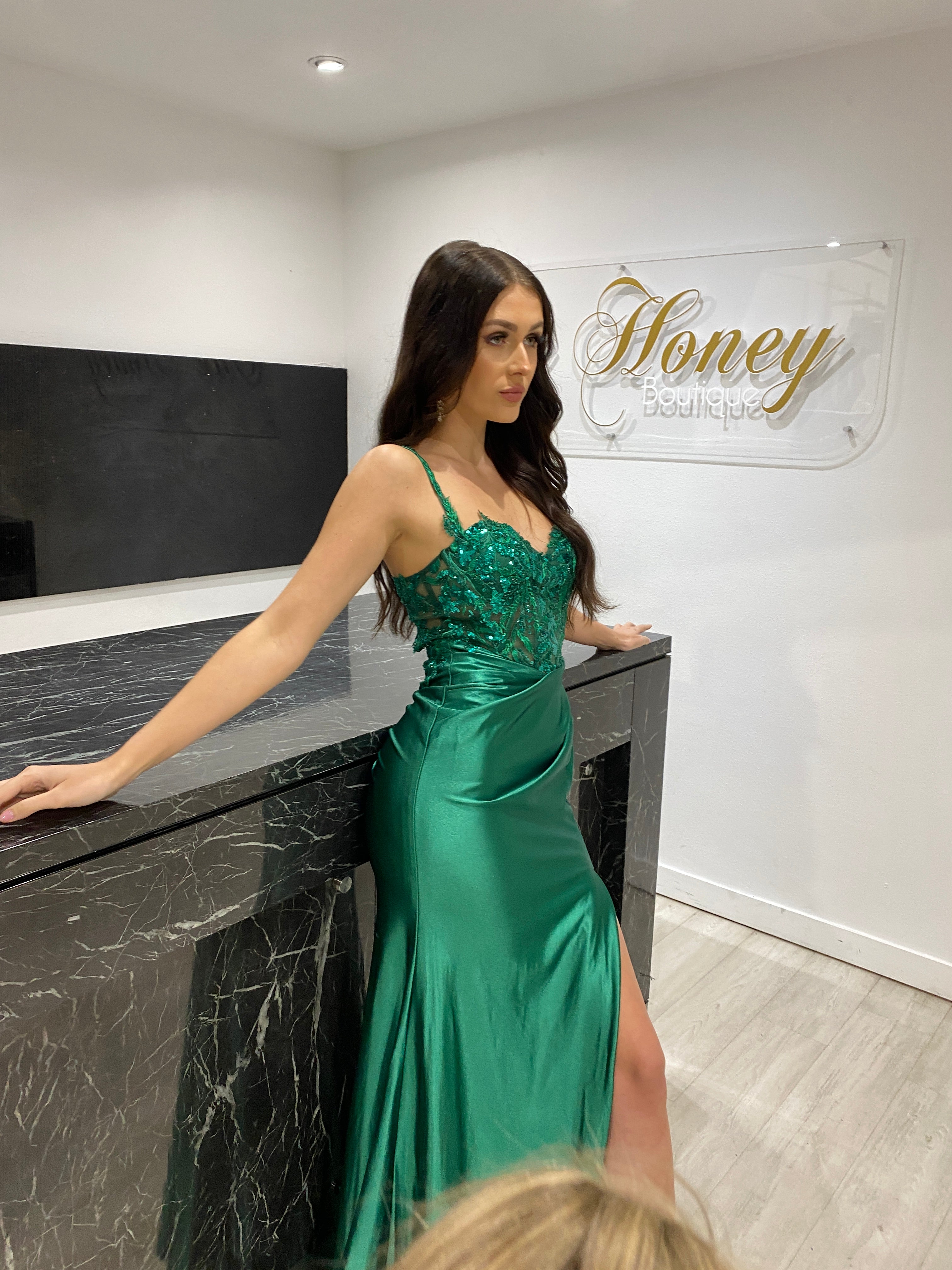 Woman in green satin mermaid formal dress with lace bodice and thin straps. Elegant evening gown in boutique interior setting.