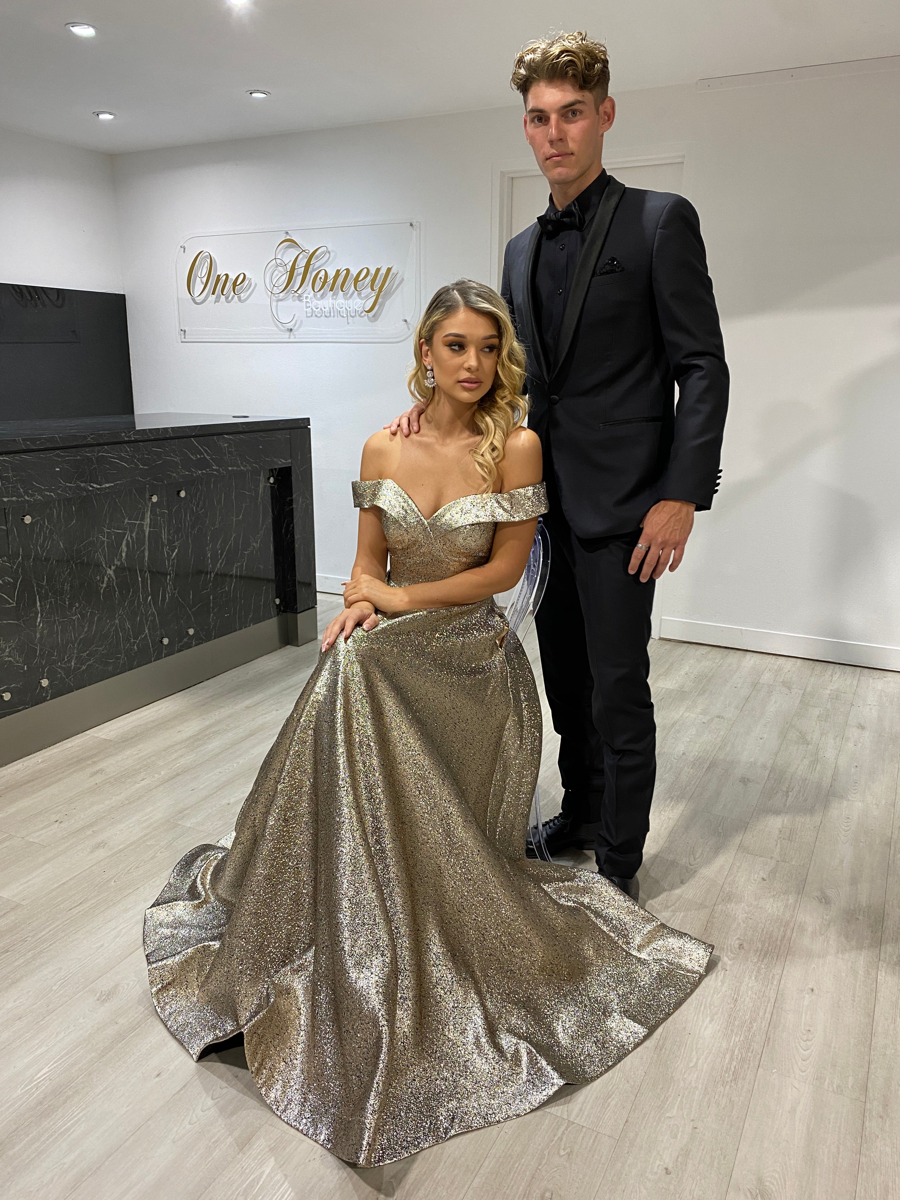 Elegant couple with woman in Honey Couture DORA bronze metallic shimmer off-shoulder gown, highlighting Australian designer prom dresses.
